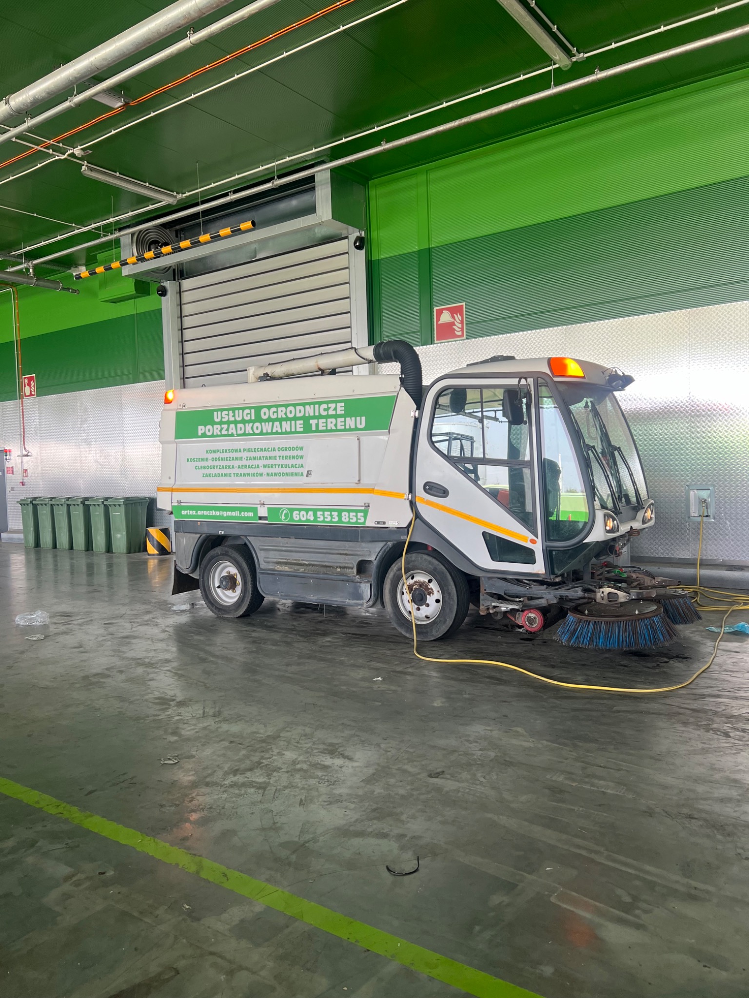 Maszyna do zamiatania z napisem 'Usługi ogrodnicze porządkowanie terenu' stoi w hali, widoczne szczotki zamiatające i pojemniki na śmieci w tle.