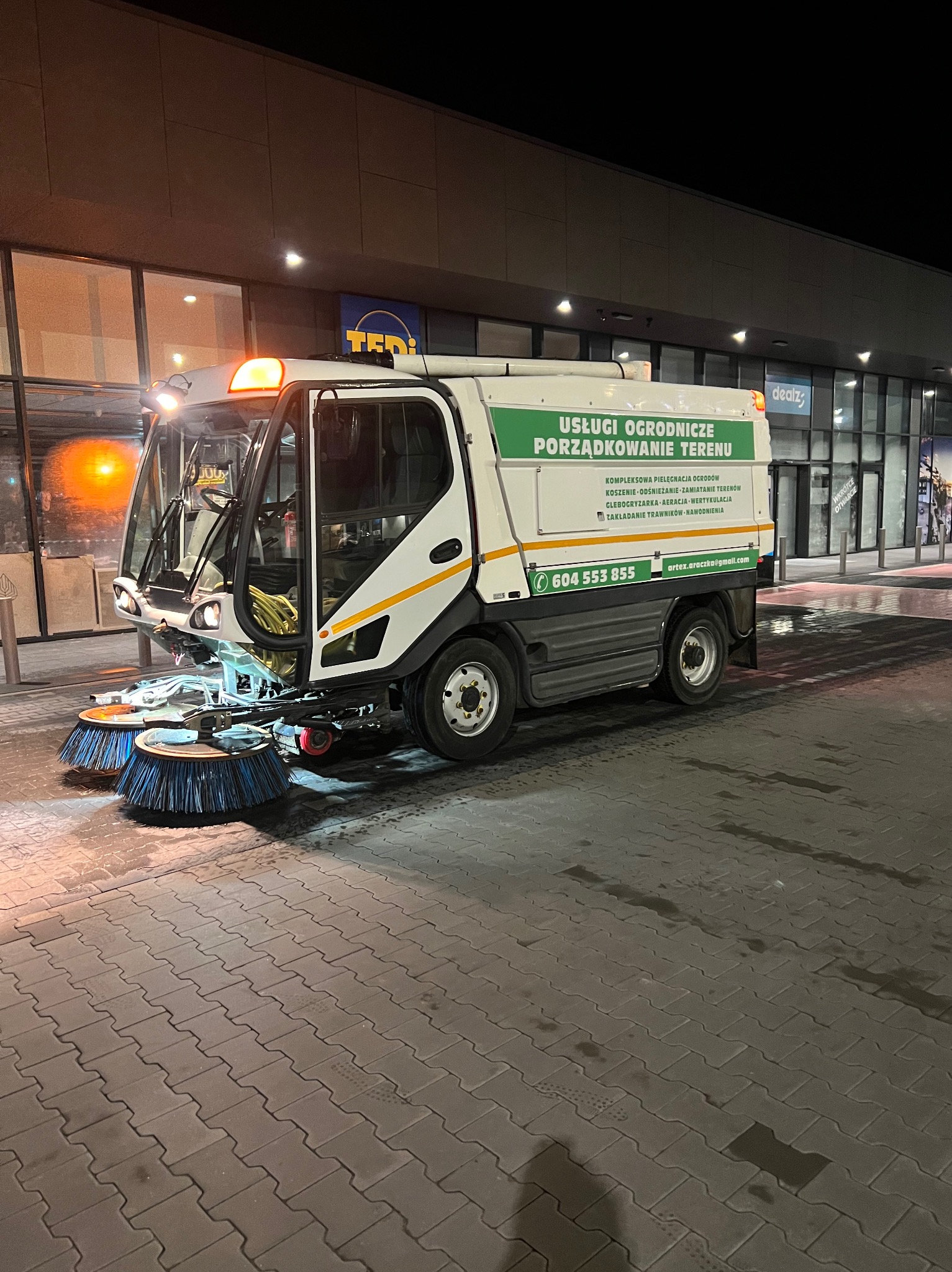 Zamiatarka uliczna z napisem 'Usługi ogrodnicze, porządkowanie terenu' zaparkowana na placu przed budynkiem sklepu w nocy.