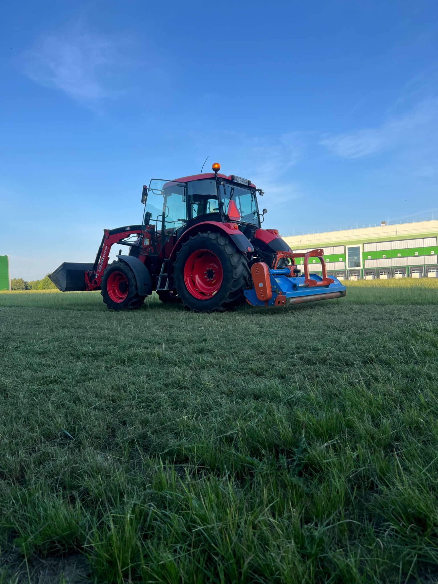 Czerwony traktor z kosiarką rotacyjną na trawiastym terenie, widoczny budynek magazynowy w tle.