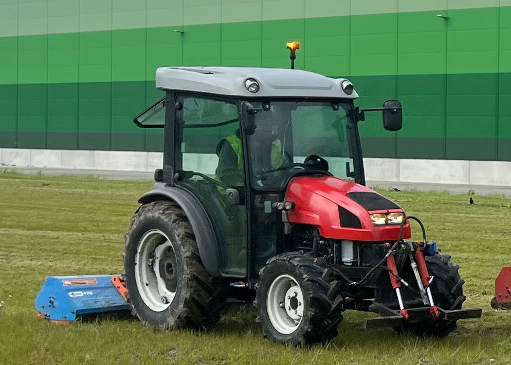 Czerwony traktor z kabiną i zamontowaną kosiarką rotacyjną na trawniku przed nowoczesnym budynkiem w odcieniach zieleni.