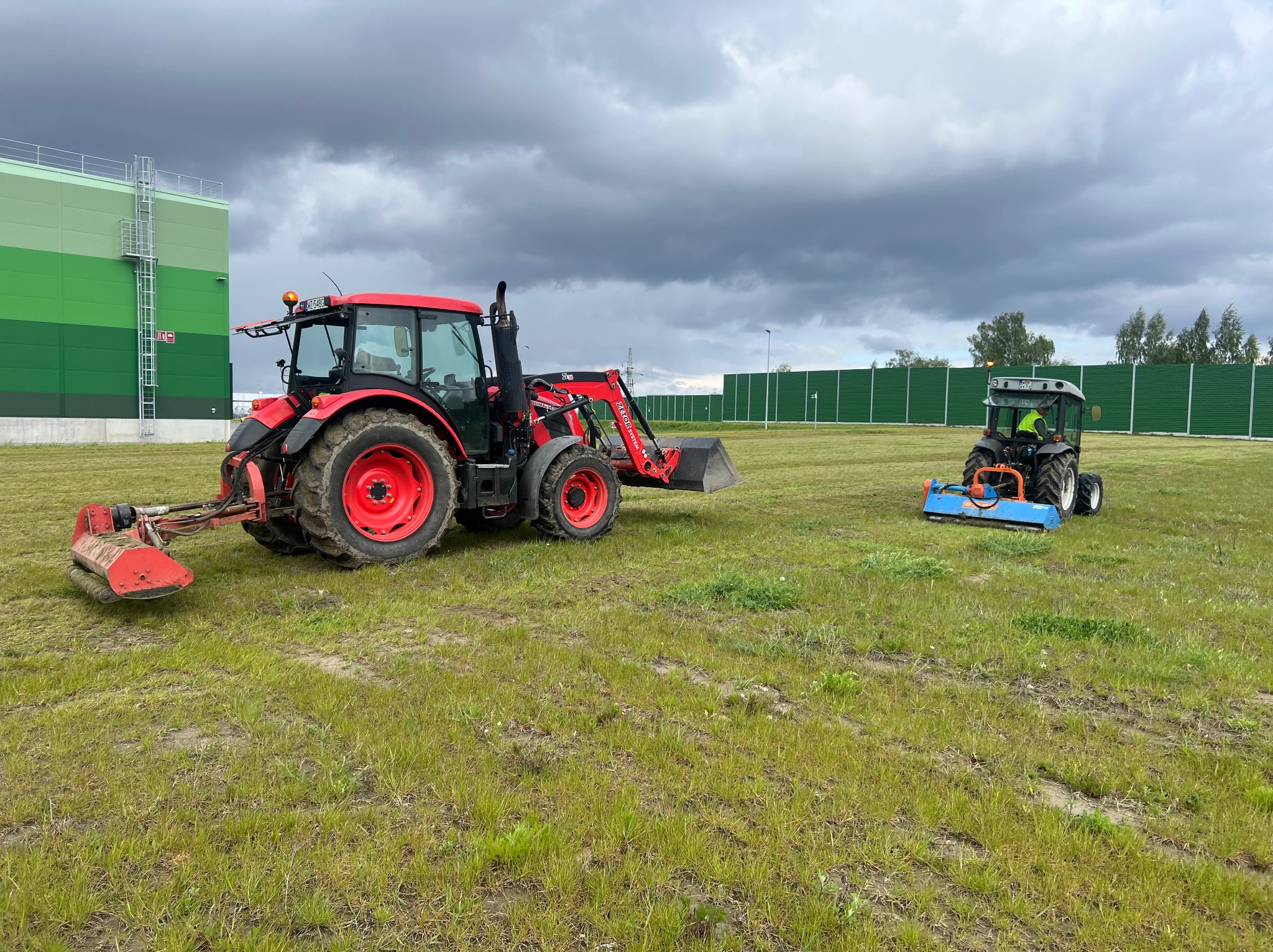 Dwa traktory koszą trawę na dużym terenie, w tle budynek przemysłowy i ekran akustyczny, pochmurne niebo.