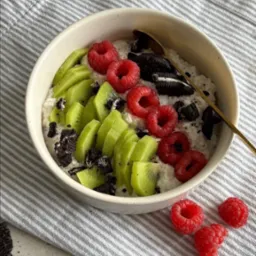 Owsianka w miseczce udekorowana plastrami kiwi, malinami i pokruszonymi ciastkami Oreo, ułożona na ściereczce w szaro-białe paski z napisem 'Enjoy life, it's delicious!'