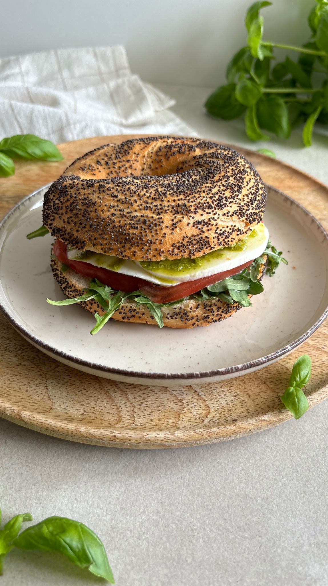 Bagel z makiem wypełniony rukolą, plastrem pomidora, mozzarellą i pesto, podany na talerzu na drewnianej tacy, z listkami bazylii w tle.