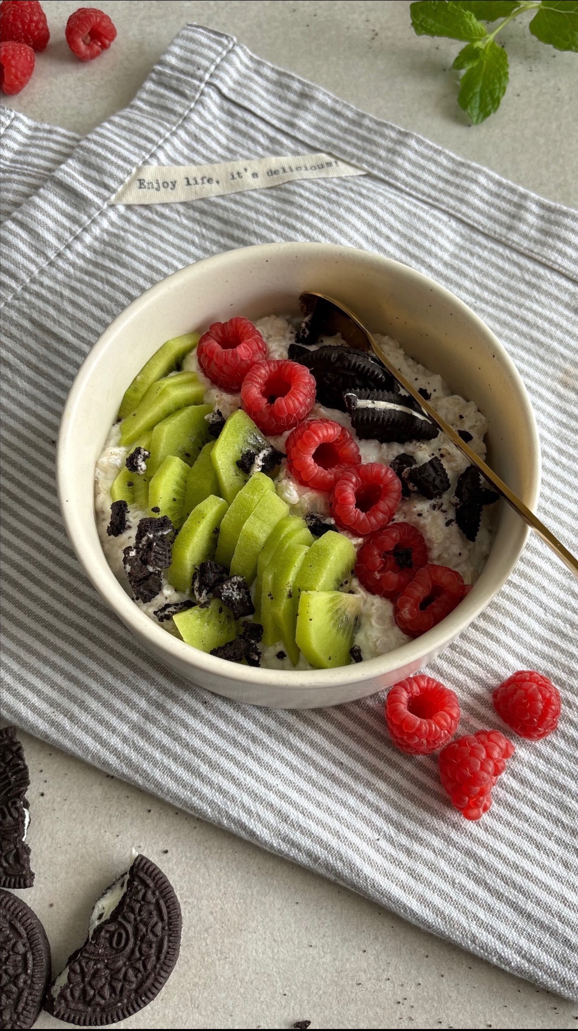 Owsianka w miseczce udekorowana plastrami kiwi, malinami i pokruszonymi ciastkami Oreo, ułożona na ściereczce w szaro-białe paski z napisem 'Enjoy life, it's delicious!'