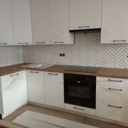 KRZYSZTOF DĄBROWSKI WYKOŃCZENIA WNĘTRZ - Nowoczesna kuchnia z białymi szafkami, drewnianym blatem i białymi płytkami ułożonymi w jodełkę na ścianie, jeszcze z folią ochronną na frontach szafek.