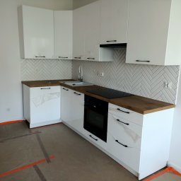 KRZYSZTOF DĄBROWSKI WYKOŃCZENIA WNĘTRZ - Nowoczesna kuchnia z białymi szafkami, drewnianym blatem i białymi płytkami ułożonymi w jodełkę, zabezpieczona folią ochronną na frontach szafek i podłodze.