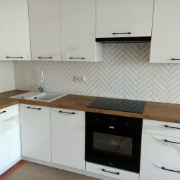 KRZYSZTOF DĄBROWSKI WYKOŃCZENIA WNĘTRZ - Nowoczesna kuchnia z białymi szafkami z czarnymi uchwytami, drewnianym blatem i białymi płytkami ułożonymi w jodełkę, częściowo jeszcze zabezpieczona folią ochronną.