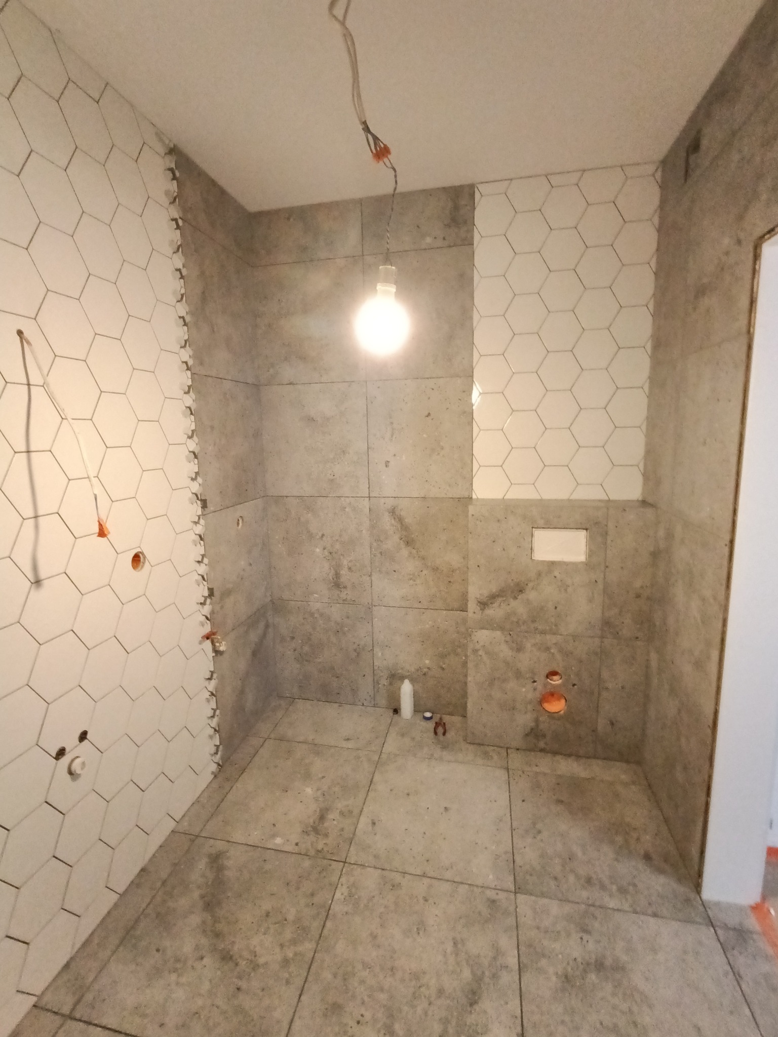 Łazienka w trakcie układania płytek: szare, kwadratowe płytki na ścianach i podłodze kontrastują z białymi, heksagonalnymi płytkami na fragmencie ściany; widoczne niedokończone krawędzie...