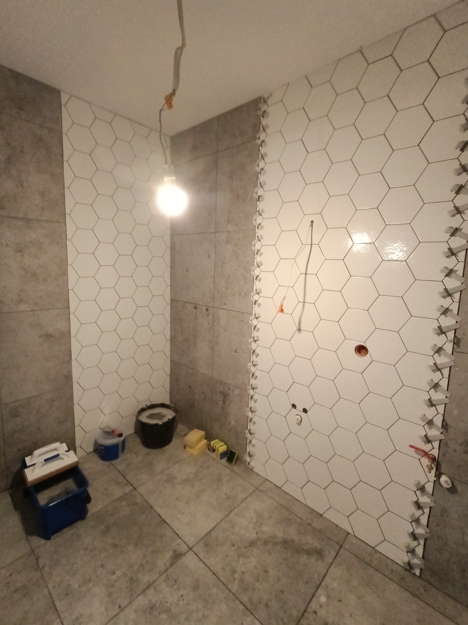 Układanie białych, heksagonalnych płytek na ścianie w łazience, widoczne krzyżyki dystansowe, szare płytki na pozostałych ścianach i podłodze, wisząca żarówka, narzędzia glazurnicze w tle.