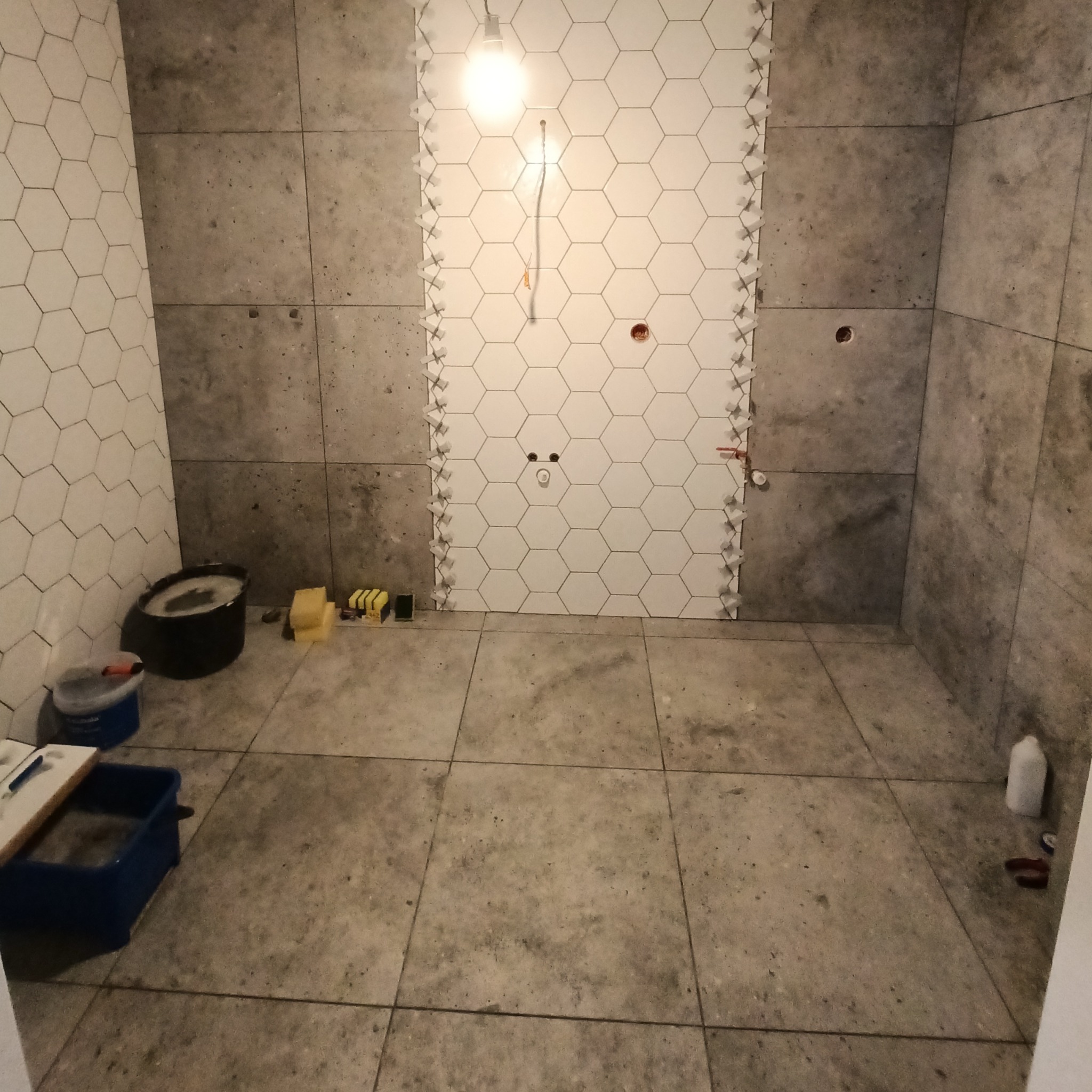 Łazienka w trakcie prac: białe, heksagonalne płytki układane na ścianie z użyciem krzyżyków dystansowych, szare płytki na podłodze i ścianach, widoczne narzędzia glazurnicze i wiadro z zaprawą.