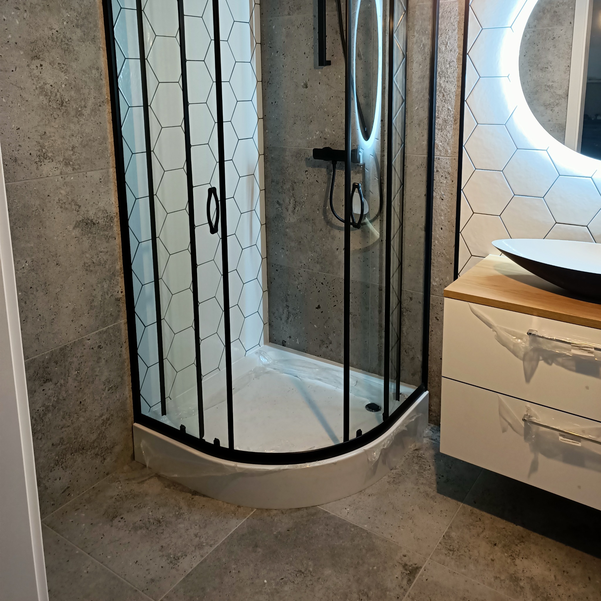 Nowoczesna kabina prysznicowa z czarnymi profilami i wzorem plastra miodu na szkle, zamontowana w łazience z szarymi płytkami i umywalką na drewnianym blacie, jeszcze zabezpieczona folią ochronną.