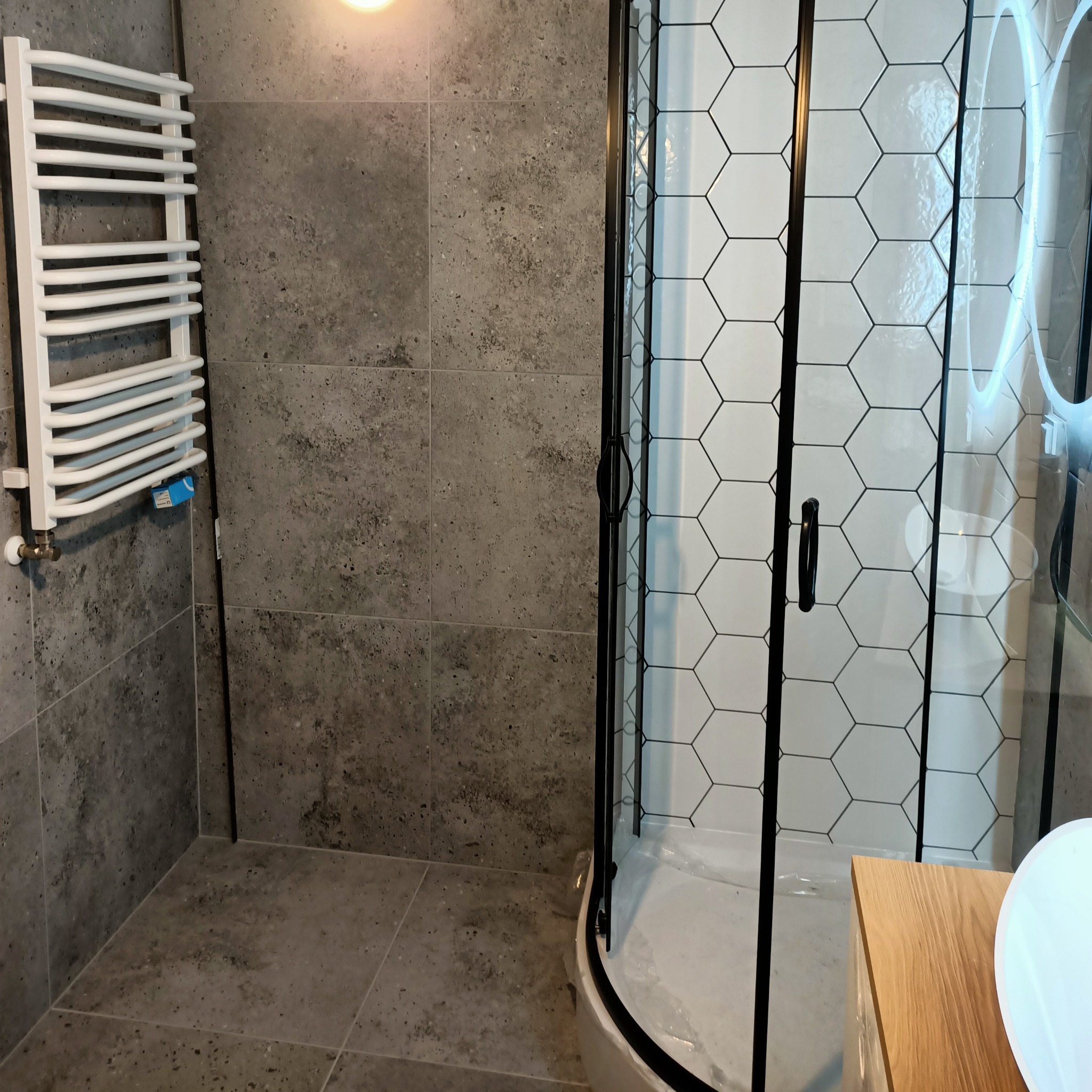 Nowoczesna łazienka z szarymi płytkami imitującymi beton, biały grzejnik drabinkowy i kabina prysznicowa z czarnymi profilami i heksagonalnymi płytkami wewnątrz.