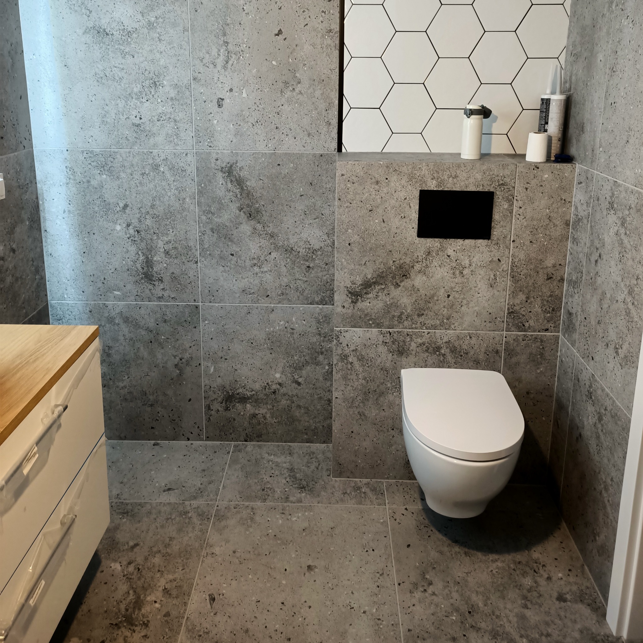 Nowoczesna łazienka z szarą, nakrapianą płytką, podwieszaną toaletą i umywalką z drewnianym blatem, akcentem heksagonalnych płytek na ścianie i czarnym przyciskiem spłukującym.