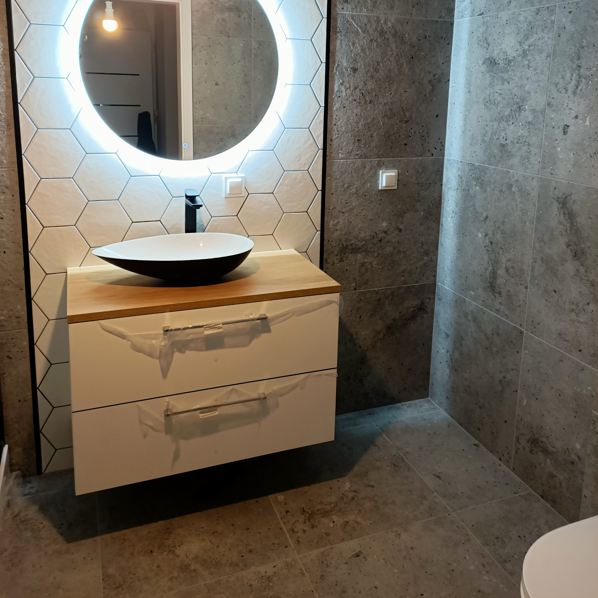 Nowoczesna łazienka z umywalką w kształcie misy, podświetlanym lustrem w kształcie koła i szarą płytką imitującą kamień, szafka pod umywalką jeszcze w folii ochronnej.