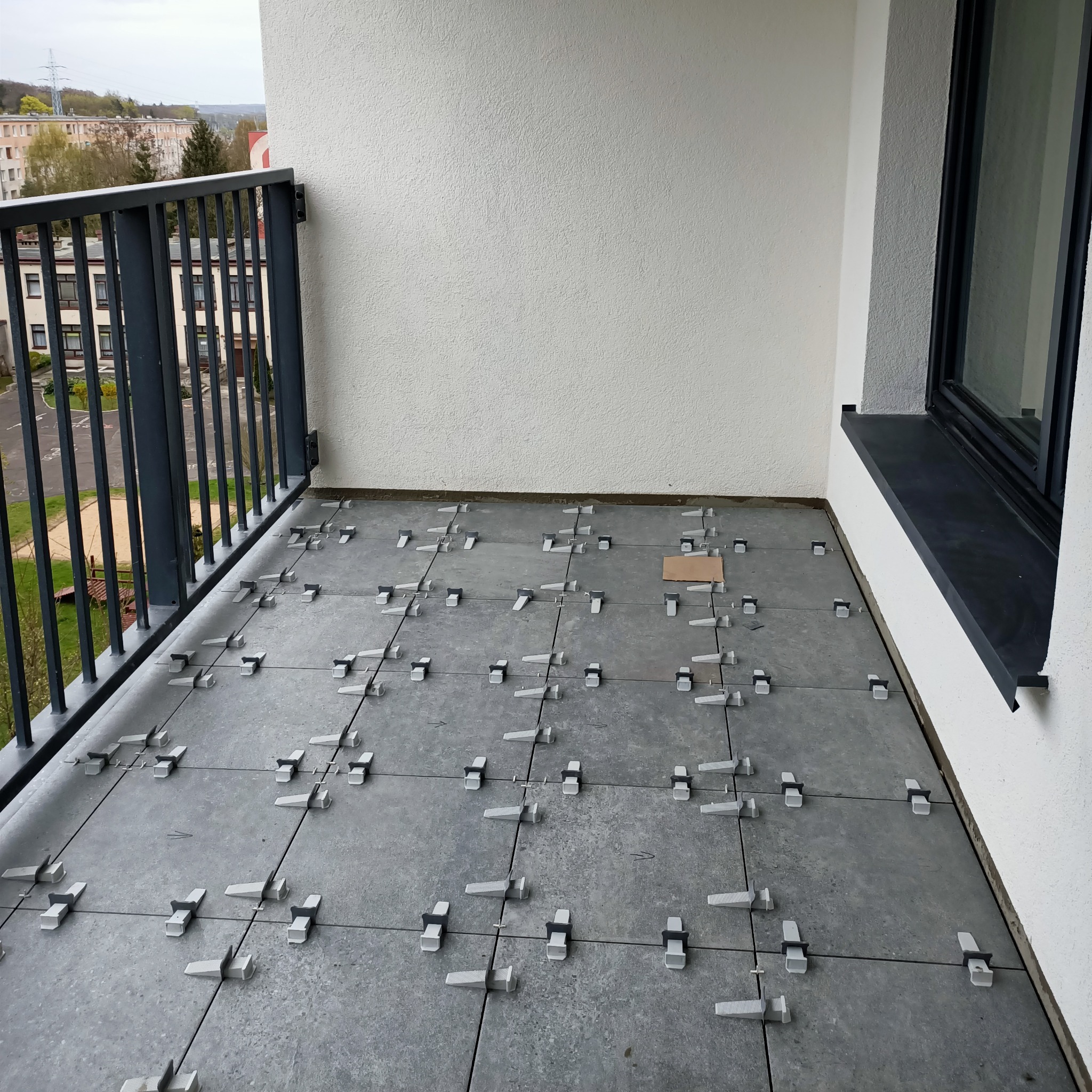 Szare płytki na balkonie w trakcie układania, z użyciem systemu poziomowania płytek i kartonowego podkładu, widok z góry na fragment balkonu z metalową barierką i oknem.