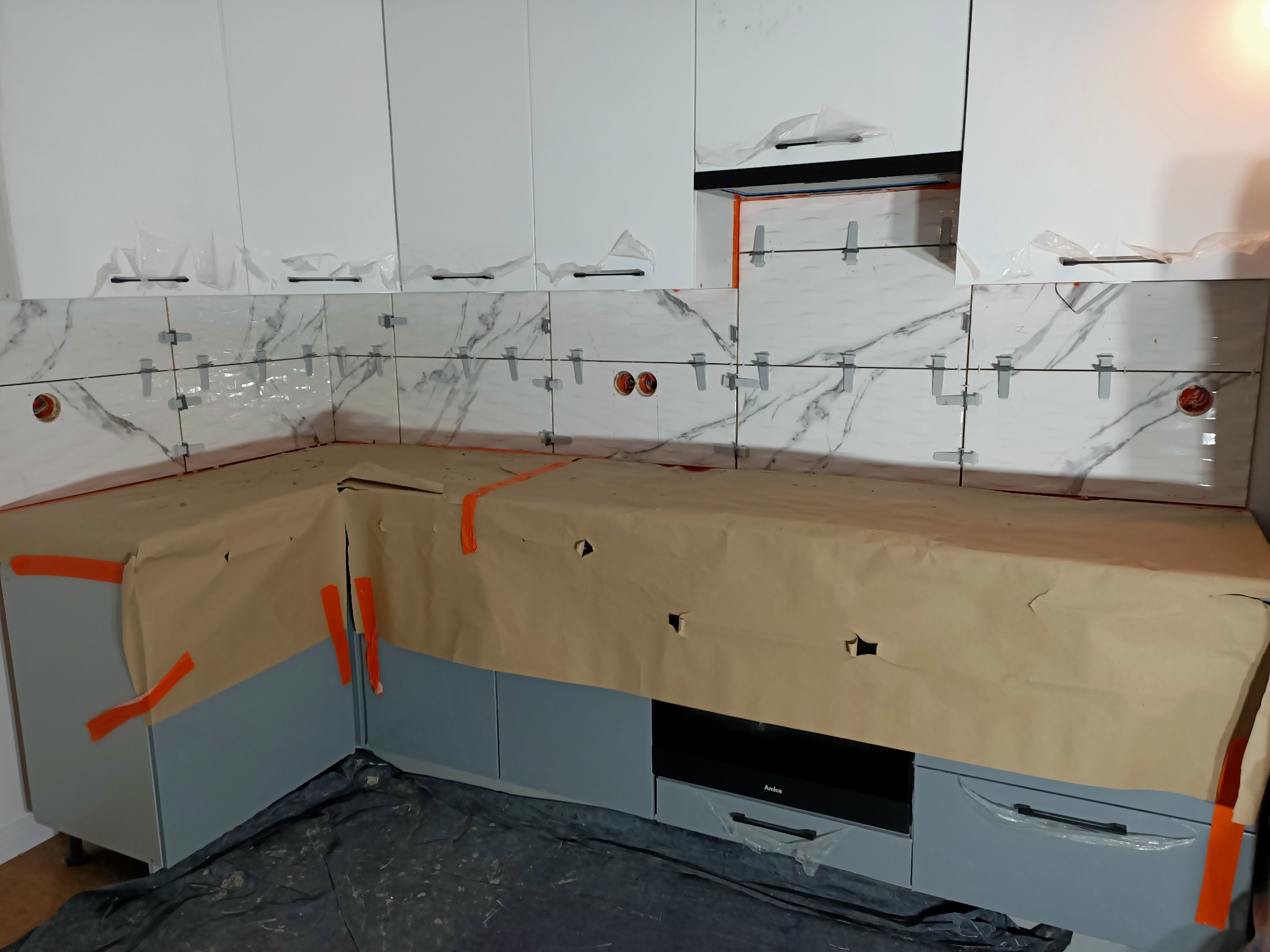 Układanie płytek imitujących marmur na ścianie w kuchni, z widocznymi krzyżykami dystansowymi i zabezpieczoną folią szafką, podczas prac wykończeniowych.