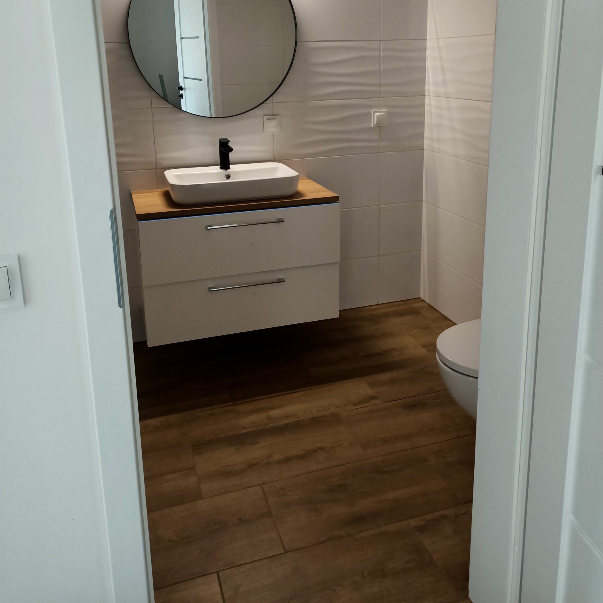 Nowoczesna łazienka z umywalką nablatową na szafce z dwiema szufladami, okrągłym lustrem i płytkami z delikatnym wzorem fali na ścianie, widok zza uchylonych białych drzwi.