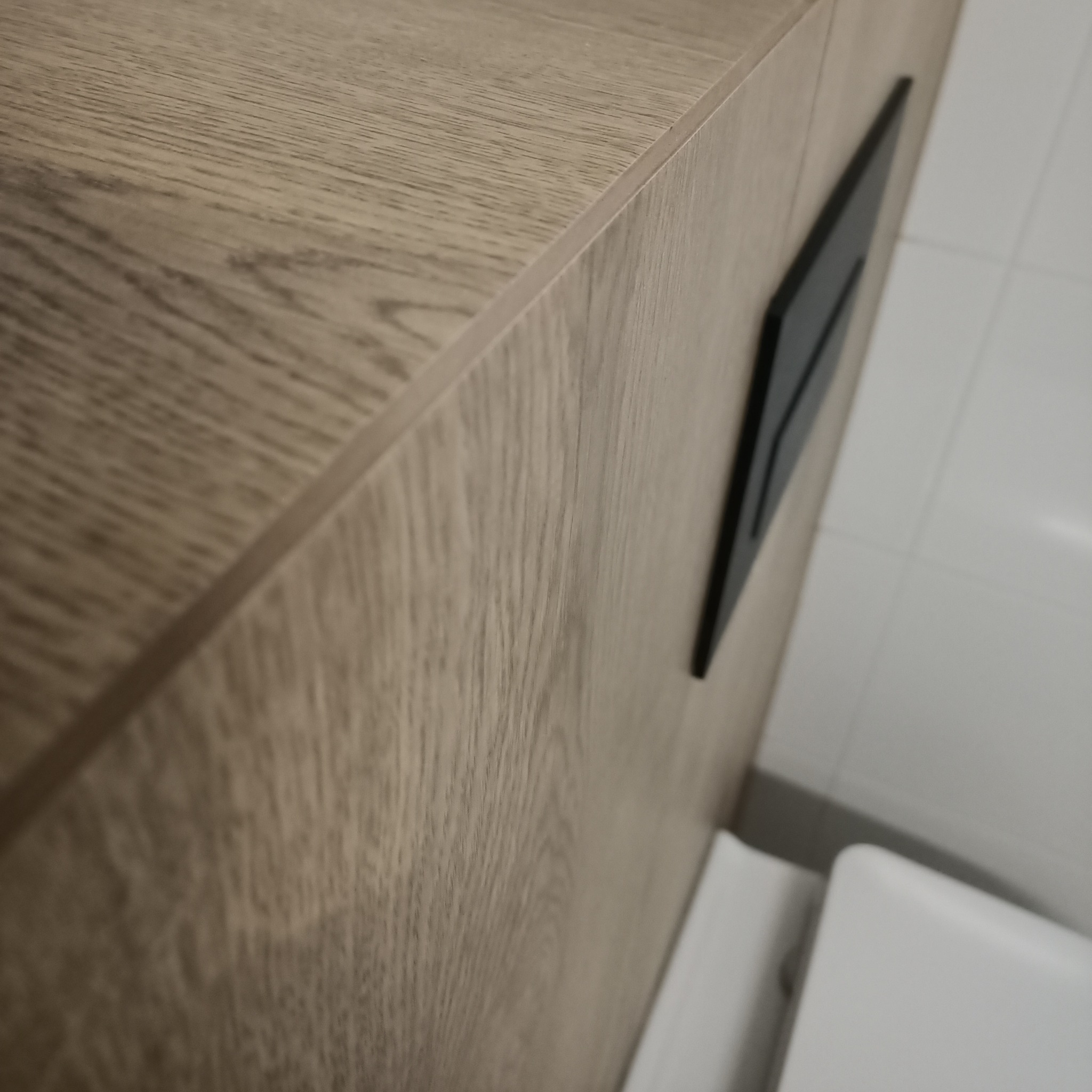 Drewnopodobna szafka łazienkowa z czarnym panelem sterującym spłuczką, widoczna biała toaleta i fragment płytek ściennych.