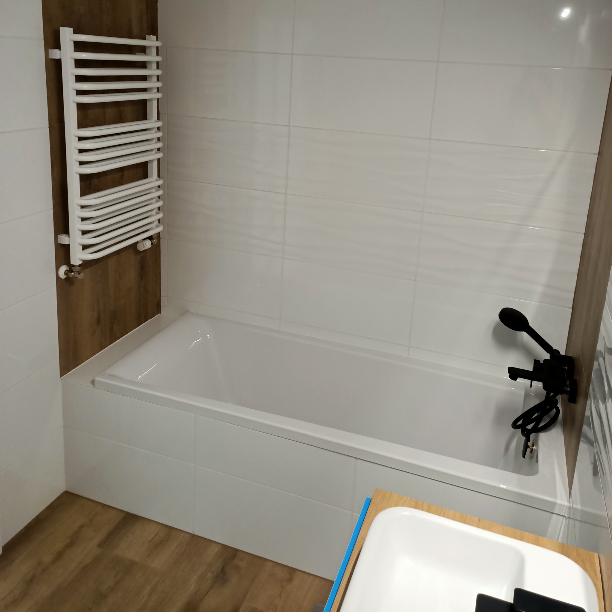 Nowoczesna łazienka z białą wanną, białymi płytkami imitującymi fale, grzejnikiem drabinkowym i czarną armaturą.