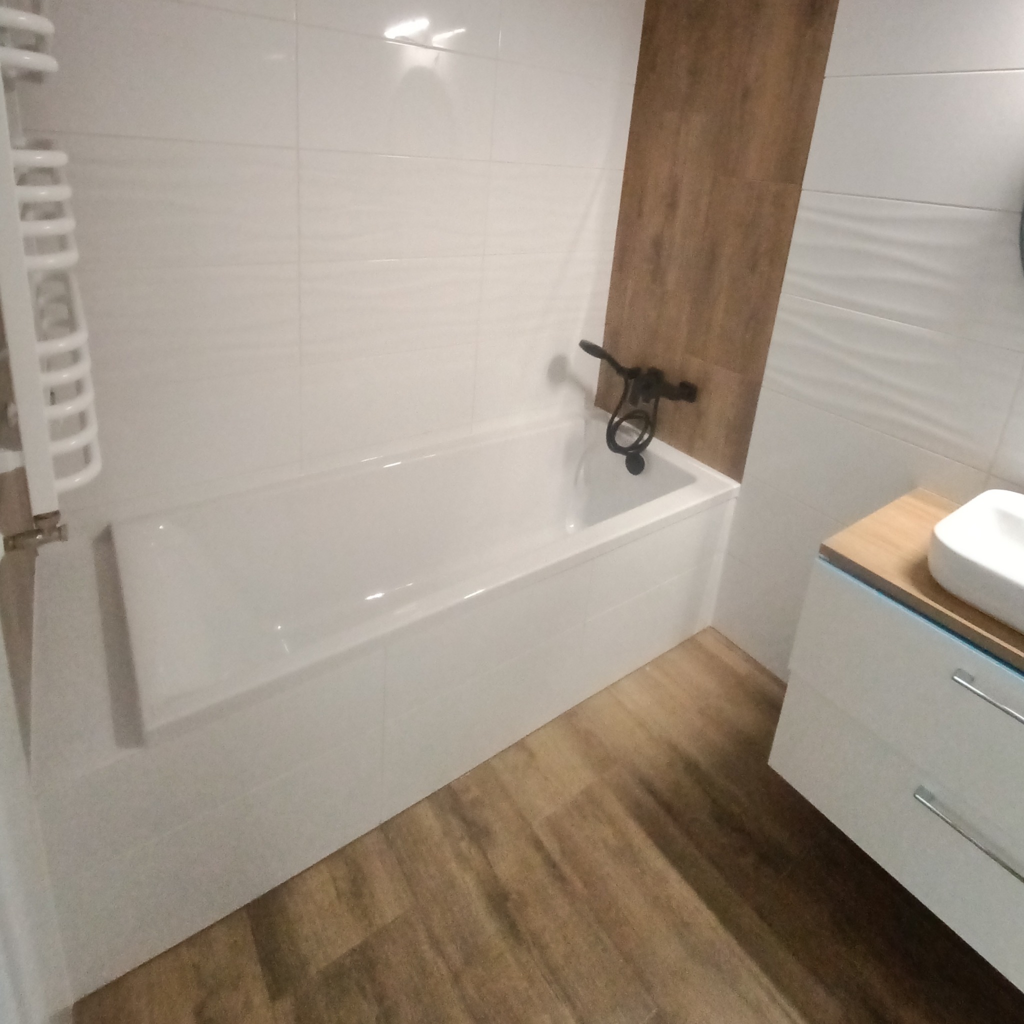 Biała wanna obudowana płytkami, czarna bateria wannowa, białe płytki ścienne z delikatną fakturą, drewnopodobne płytki na ścianie i podłodze, biała szafka pod umywalkę z drewnianym blatem.