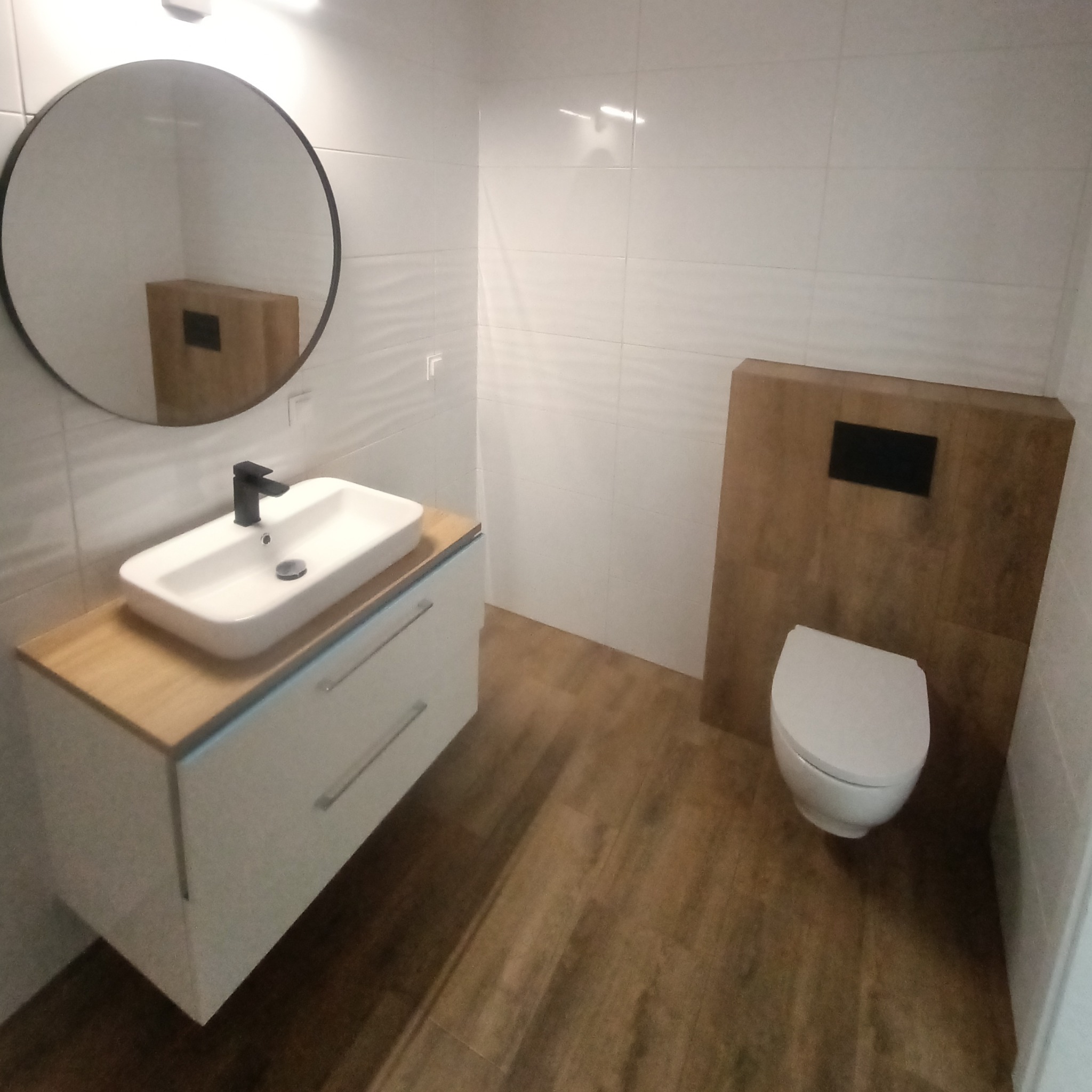 Nowoczesna łazienka z okrągłym lustrem, białą umywalką na drewnianym blacie, wiszącą toaletą i płytkami imitującymi drewno na podłodze.