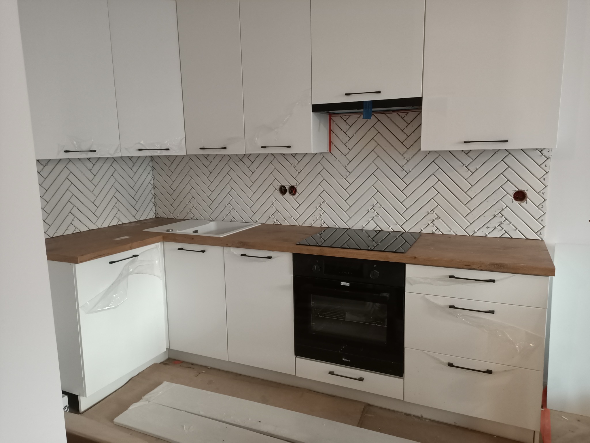 Nowoczesna kuchnia z białymi szafkami, drewnianym blatem i białymi płytkami ułożonymi w jodełkę na ścianie, jeszcze z folią ochronną na frontach szafek.