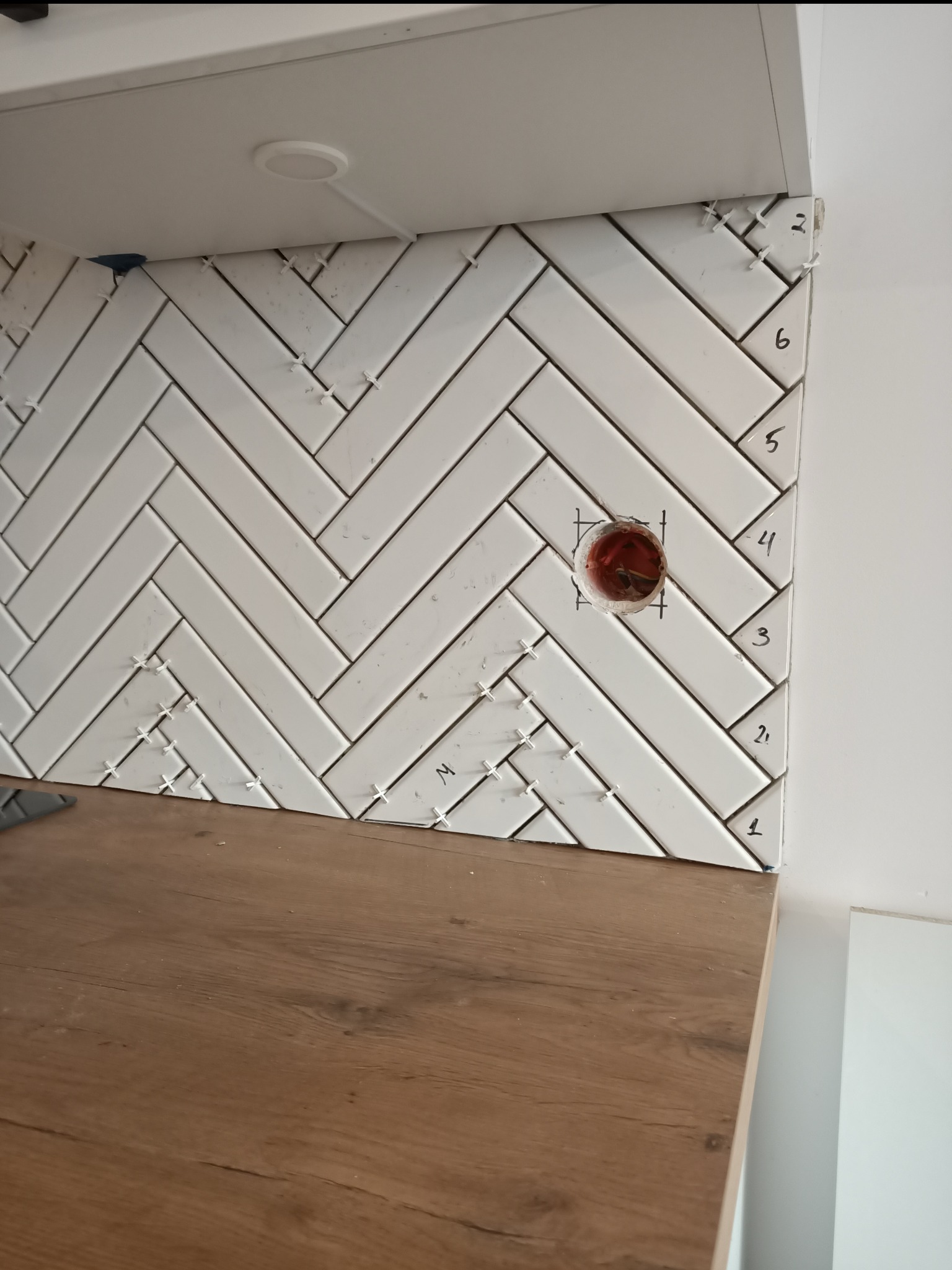 Białe, prostokątne płytki ułożone w jodełkę na ścianie nad blatem kuchennym, z widocznymi krzyżykami dystansowymi i zaznaczonym otworem na gniazdko elektryczne.