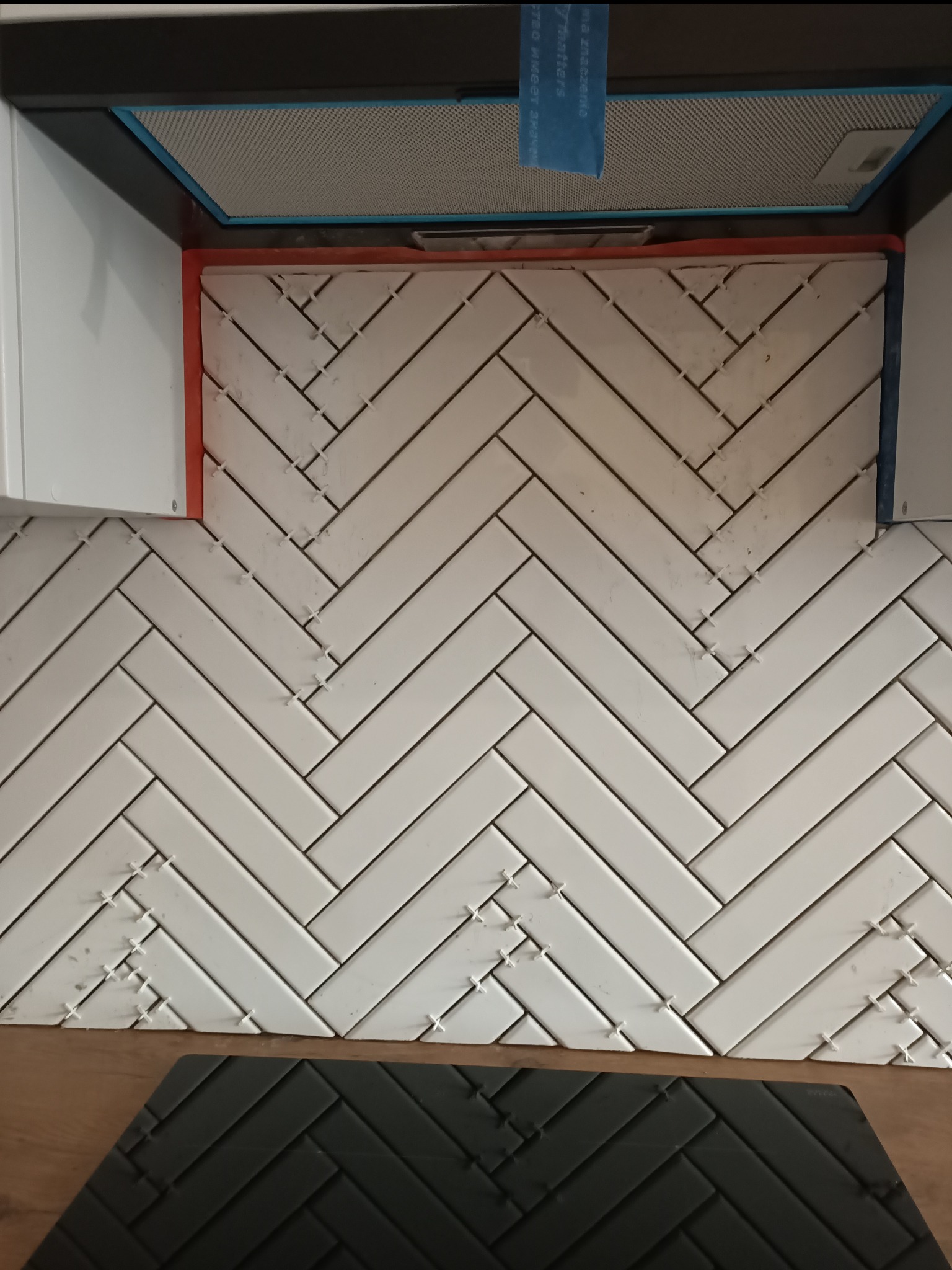 Układanie białych, prostokątnych płytek w jodełkę na ścianie nad blatem kuchennym, widoczne krzyżyki dystansowe, fragment okapu kuchennego.