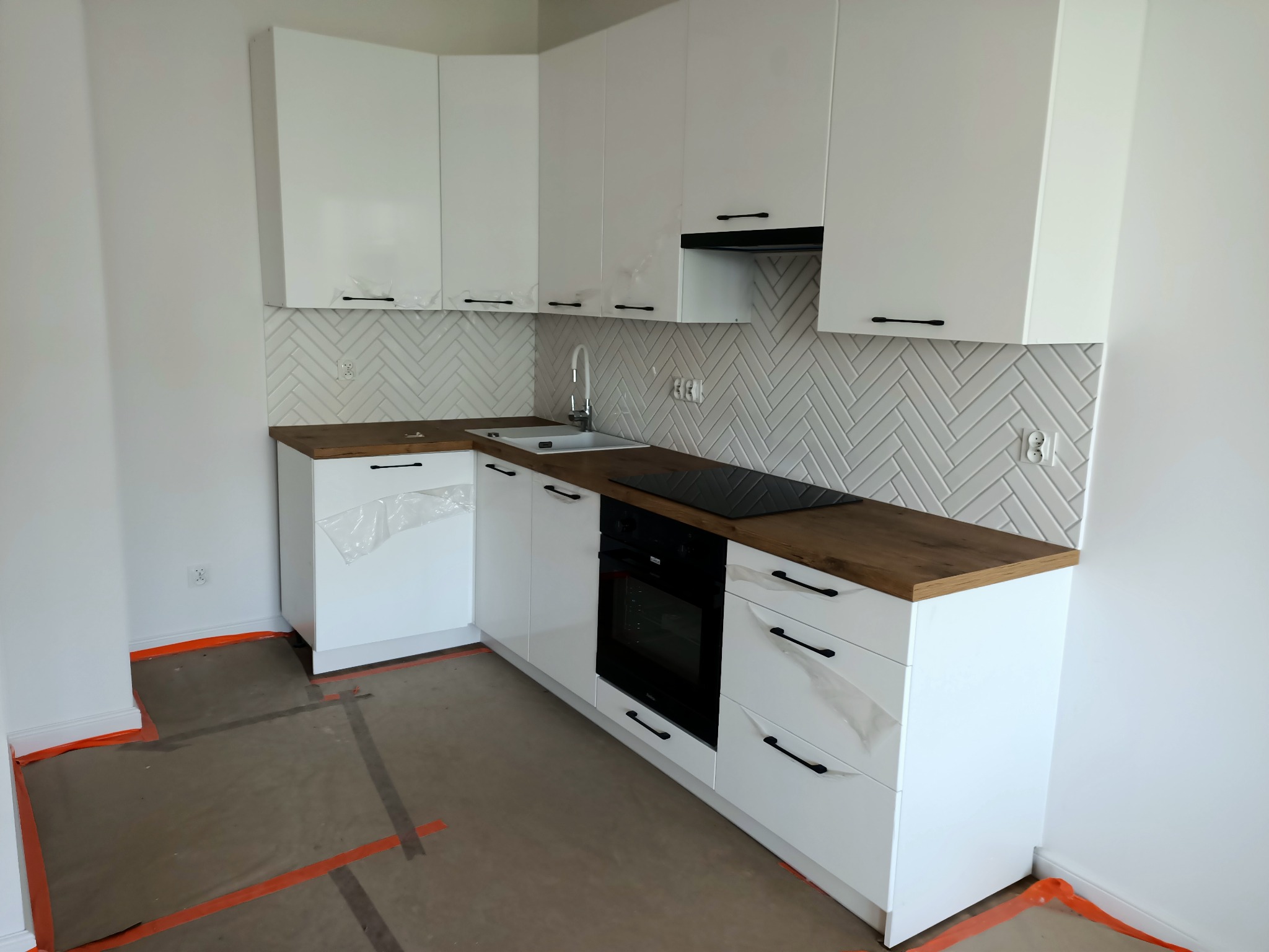 Nowoczesna kuchnia z białymi szafkami, drewnianym blatem i białymi płytkami ułożonymi w jodełkę, zabezpieczona folią ochronną na frontach szafek i podłodze.