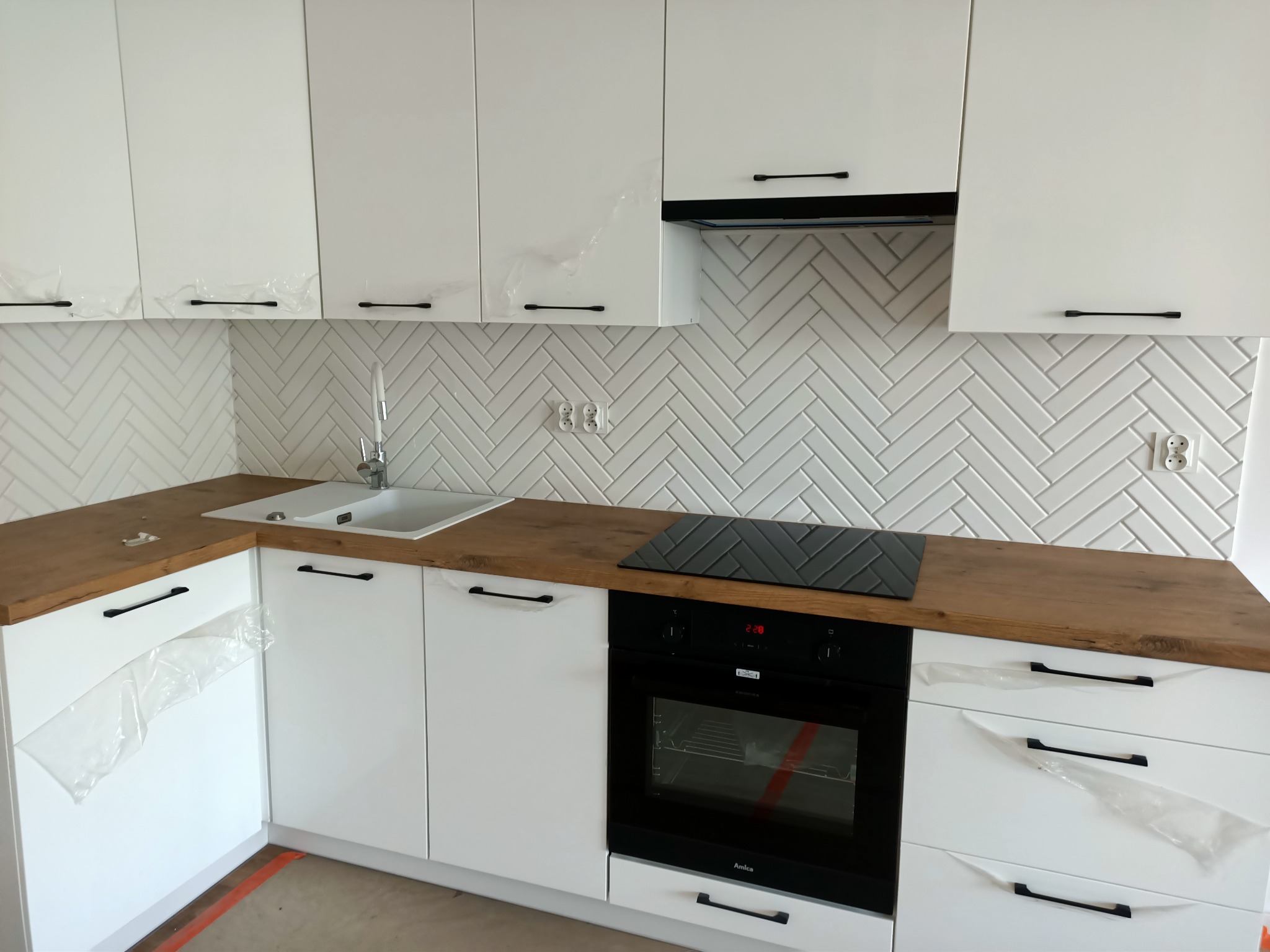 Nowoczesna kuchnia z białymi szafkami z czarnymi uchwytami, drewnianym blatem i białymi płytkami ułożonymi w jodełkę, częściowo jeszcze zabezpieczona folią ochronną.