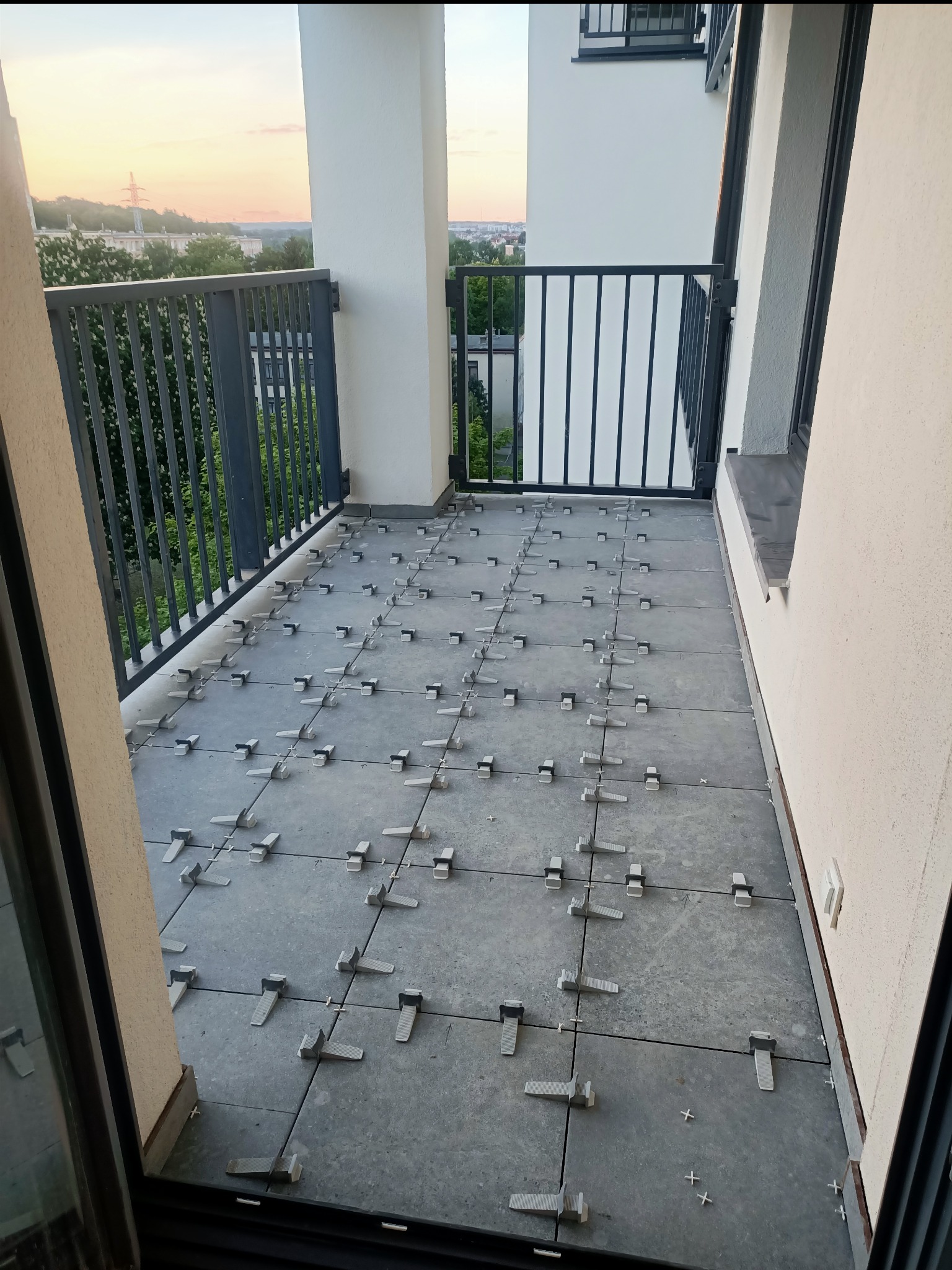 Szare płytki na balkonie w trakcie układania, z widocznymi plastikowymi klipsami poziomującymi, na tle zachodzącego słońca i miejskiego krajobrazu.