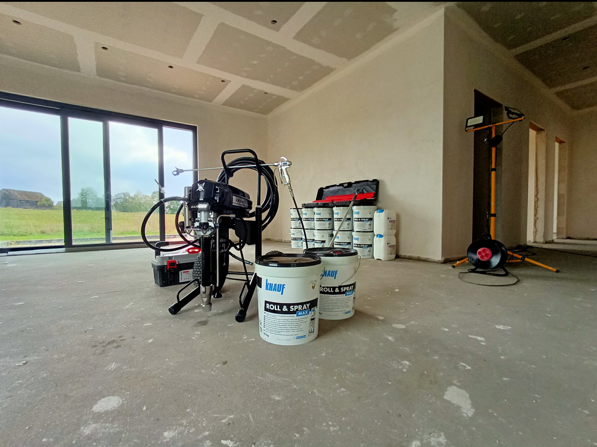 Agregat malarski Knauf Roll & Spray Max na tle surowych ścian i sufitu w trakcie szpachlowania, z widocznymi wiadrami farby i skrzynką narzędziową, oświetlony lampą budowlaną.
