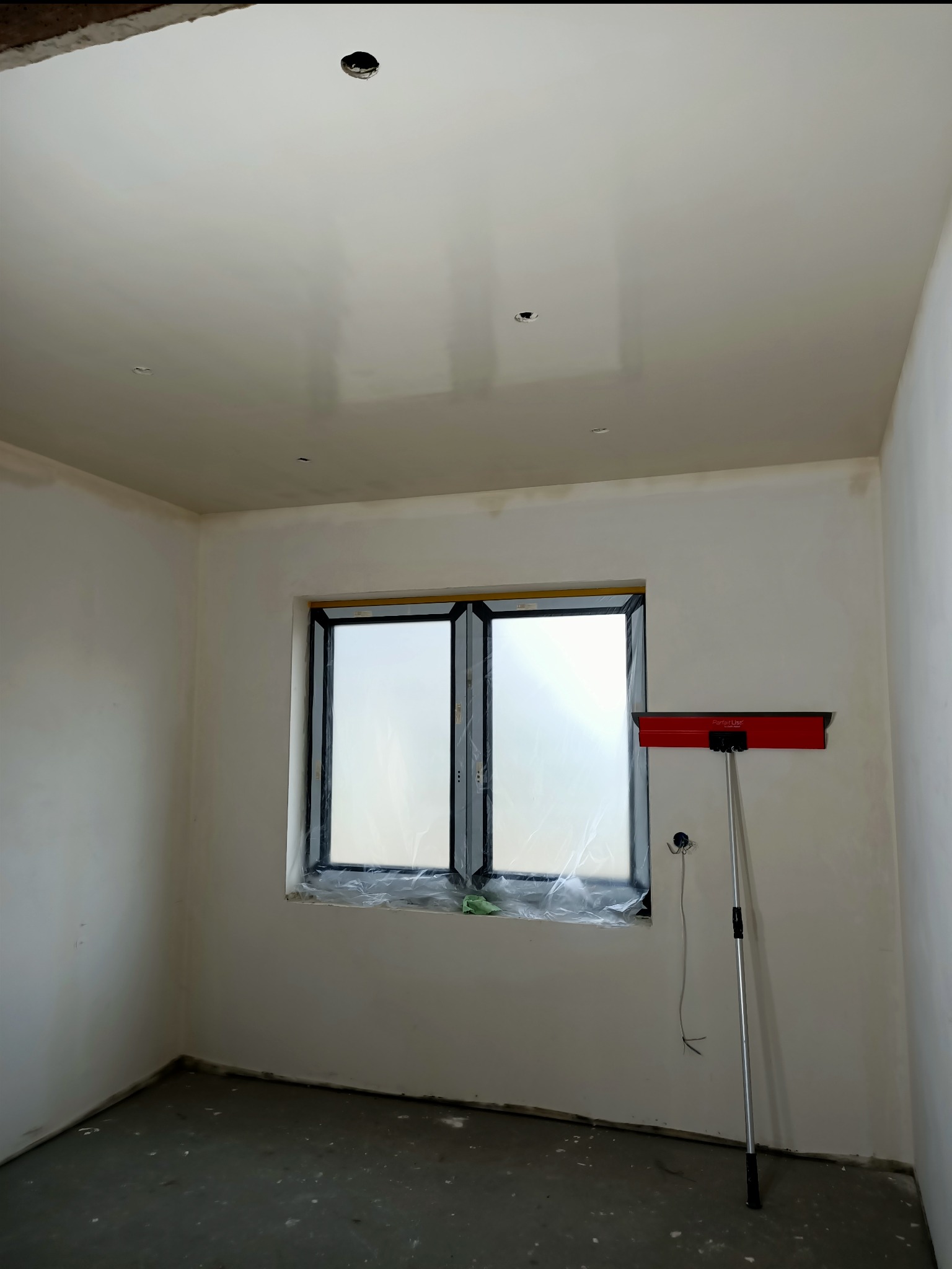 Pokój po szpachlowaniu, z widocznym oknem zabezpieczonym folią, ściany i sufit świeżo wyszpachlowane, na podłodze resztki materiałów budowlanych, oparty o ścianę długi, czerwony aplikator do szpachli.