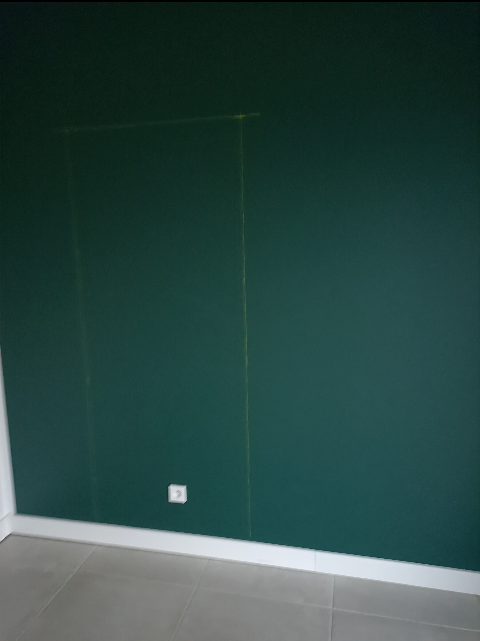 Zielona ściana z zaznaczonym ołówkiem prostokątnym obszarem, widoczne gniazdko elektryczne i szare płytki podłogowe.