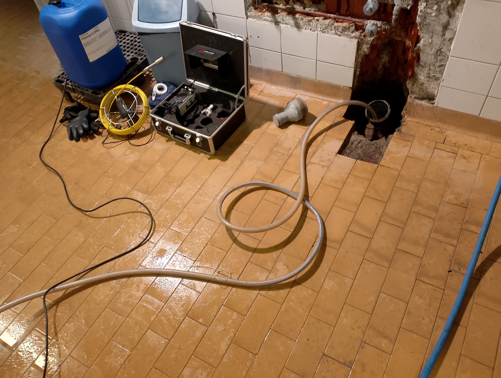 Udrażnianie rur w trakcie: kamera inspekcyjna z oświetleniem, wąż i akcesoria na mokrej, brązowej podłodze z płytek, w tle otwór w ścianie z odsłoniętymi rurami.