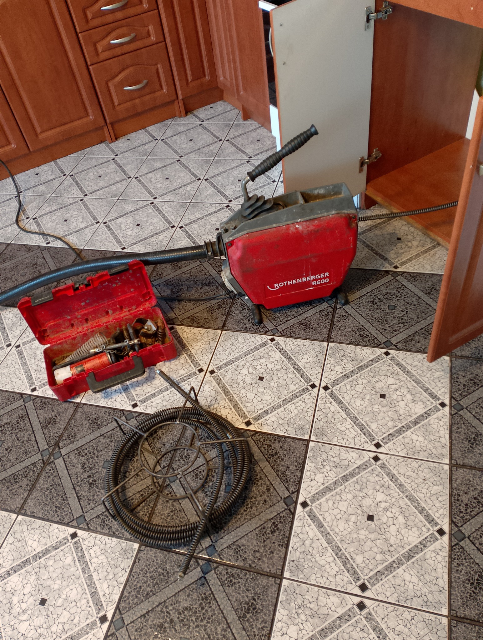 Czerwona maszyna Rothenberger R600 do przepychania rur z otwartą skrzynką narzędziową i zwiniętym wężem na wzorzystej podłodze w kuchni.