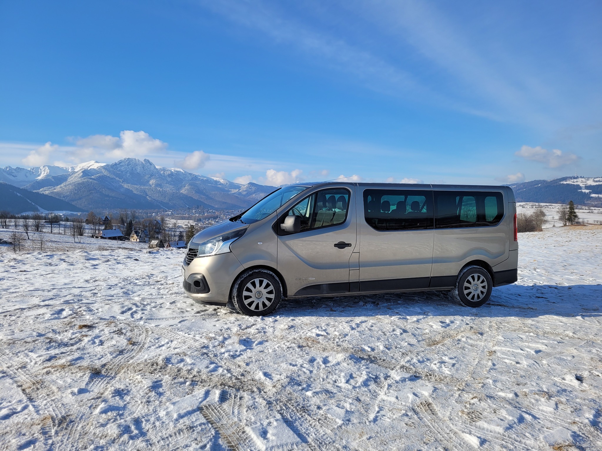 Beżowy bus Renault Trafic z przyciemnianymi szybami na śniegu z widokiem na Tatry w słoneczny dzień.