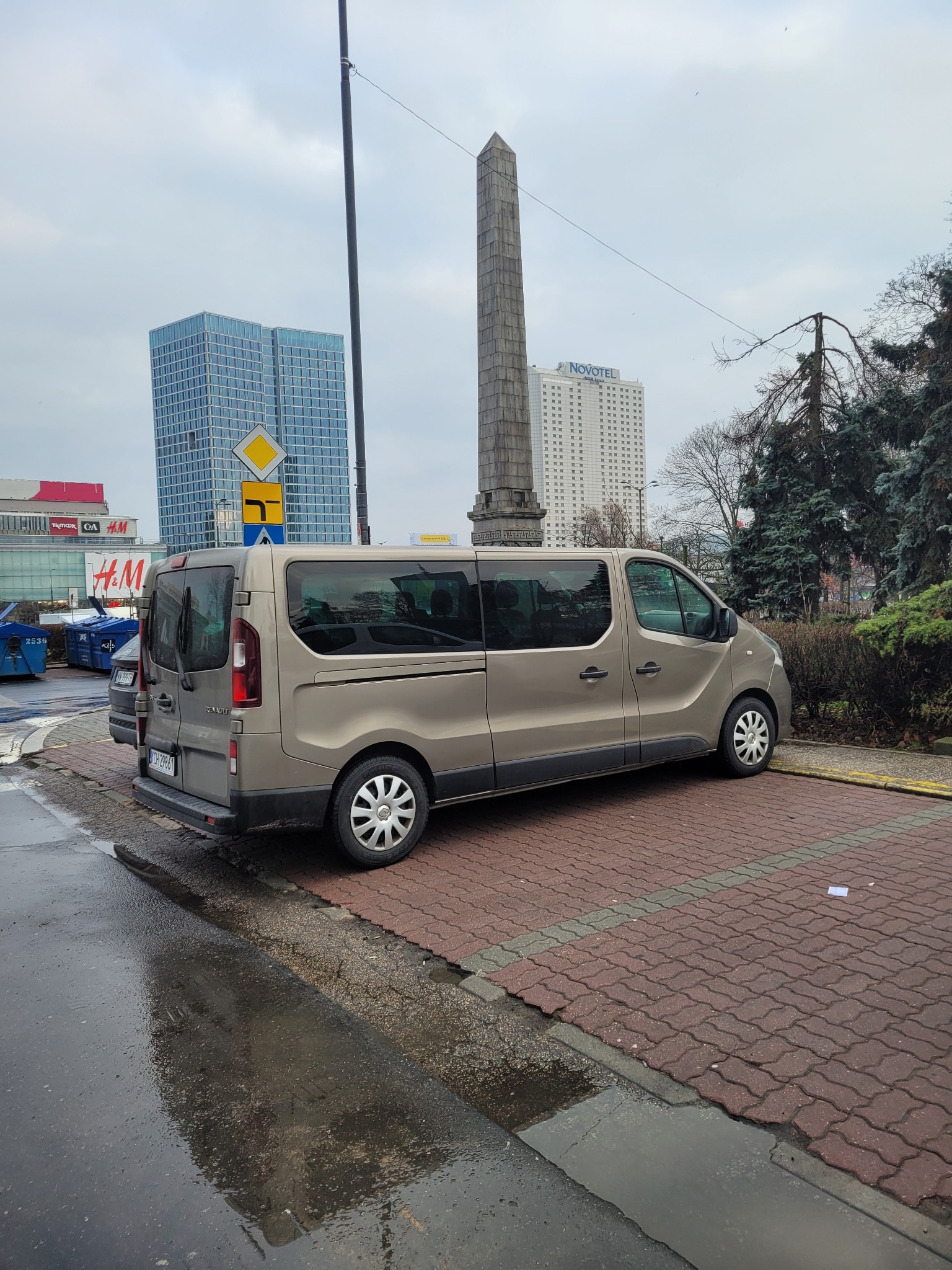 Beżowy van Renault zaparkowany na brukowanym parkingu w miejskiej scenerii, z widocznymi budynkami i pomnikiem w tle, pochmurne niebo.