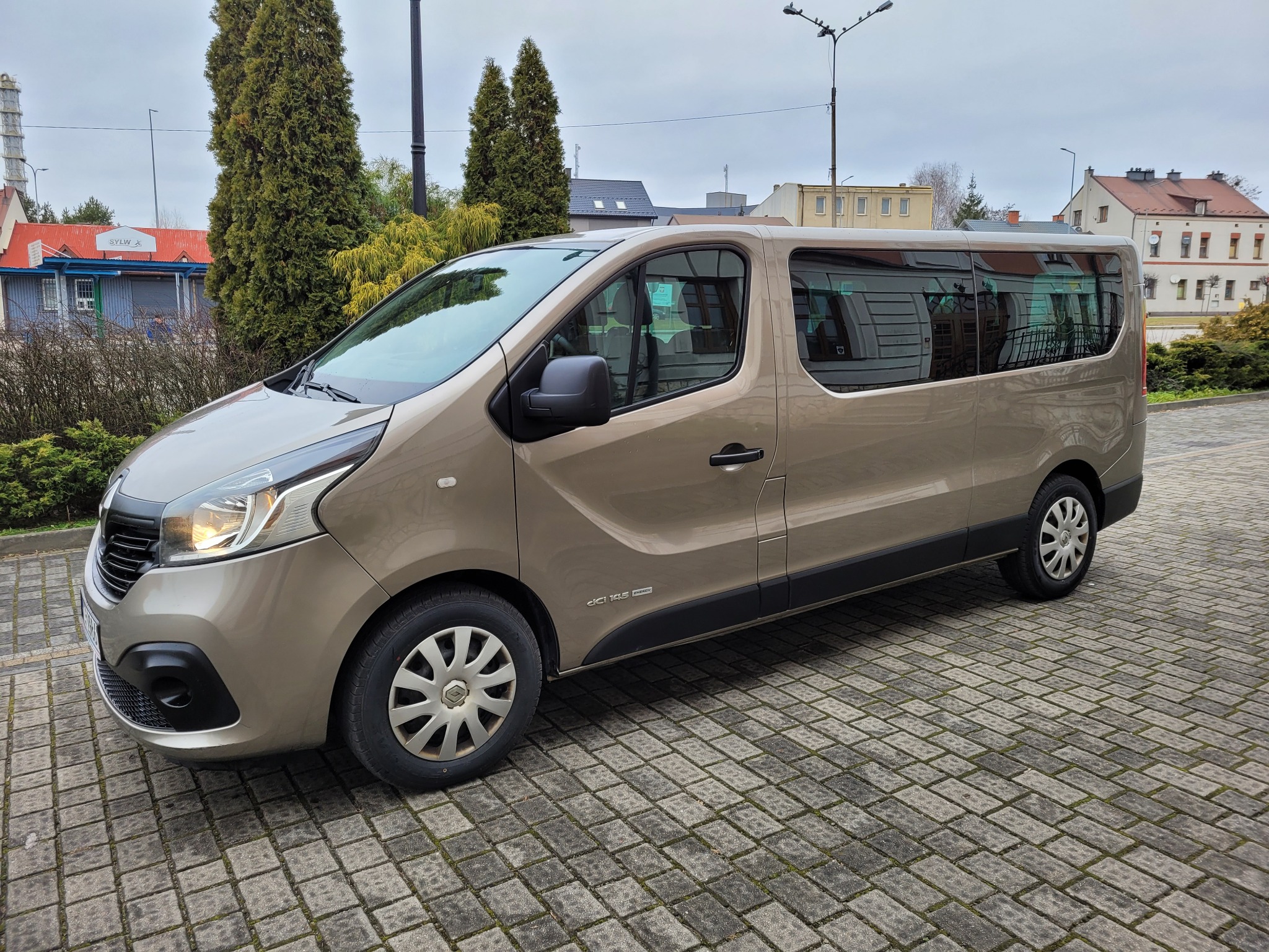 Beżowy van Renault Trafic z przyciemnianymi szybami, zaparkowany na kostce brukowej w miejskiej scenerii.