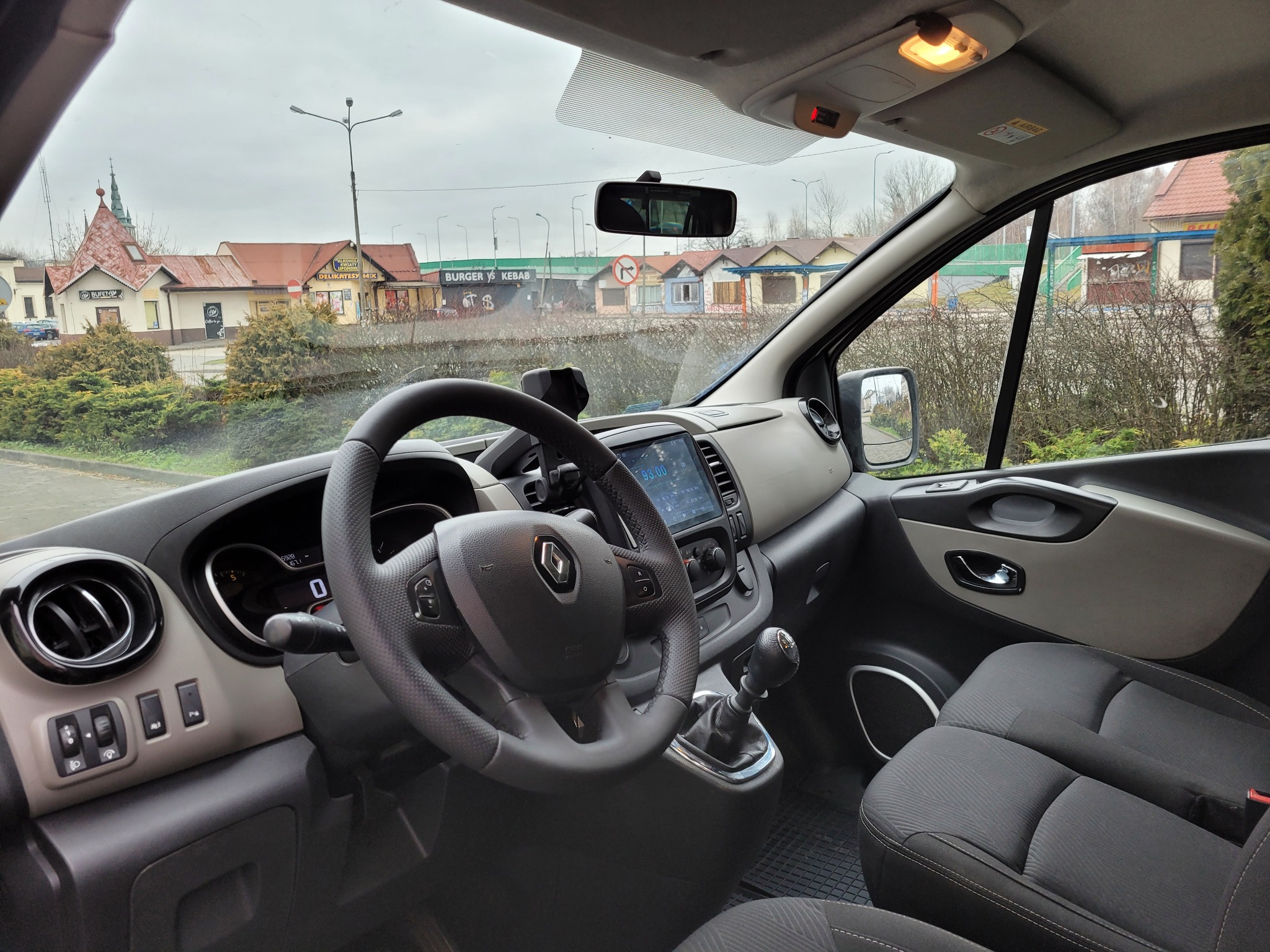 Wnętrze Renault Trafic z widokiem na zabudowania zza szyby, deska rozdzielcza w odcieniach szarości i czerni, kierownica z logo Renault.