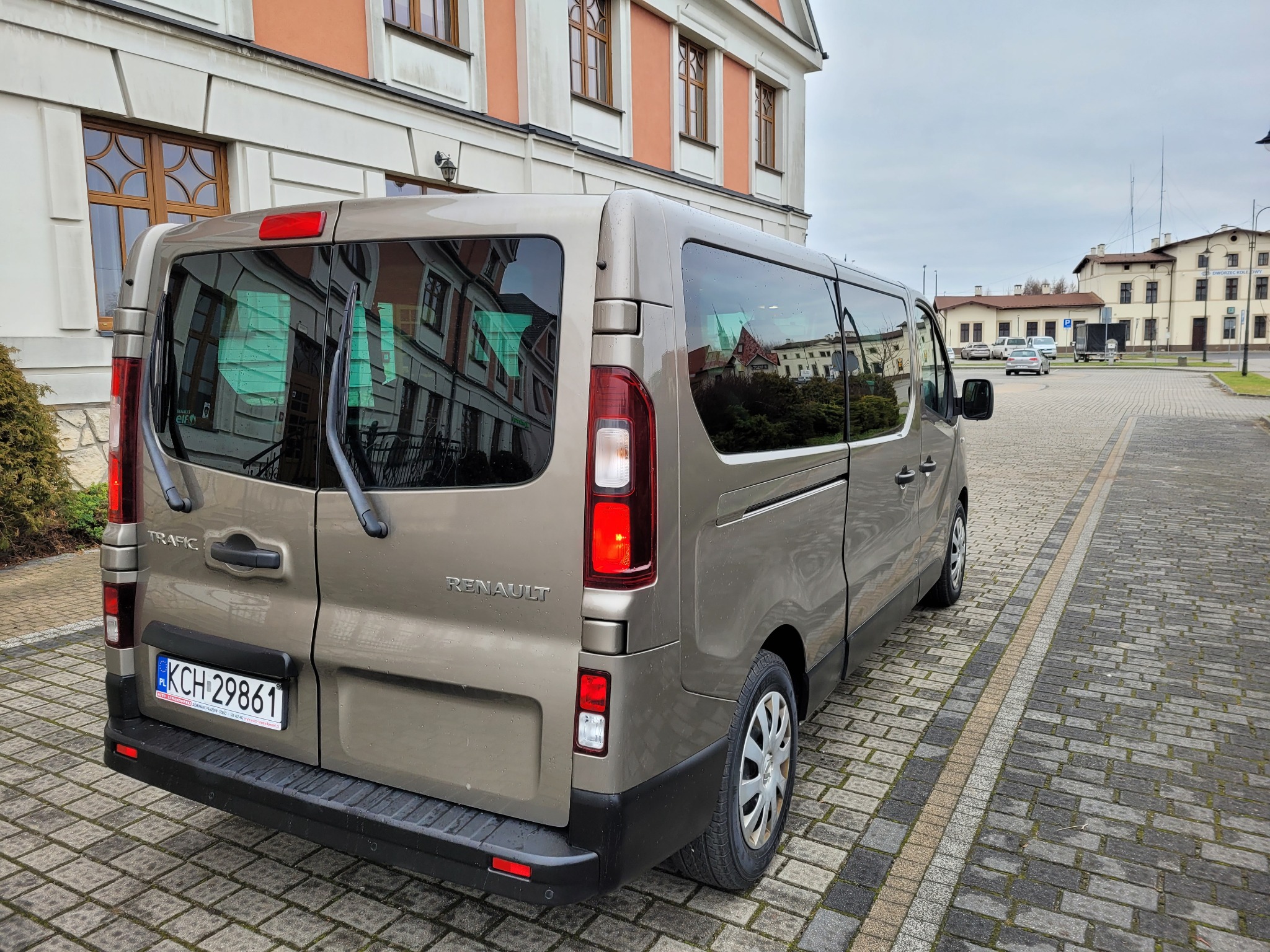 Szary Renault Trafic z numerem rejestracyjnym KCH 29861 zaparkowany na brukowanej ulicy, w tle budynek z czerwonym dachem odbity w szybach.