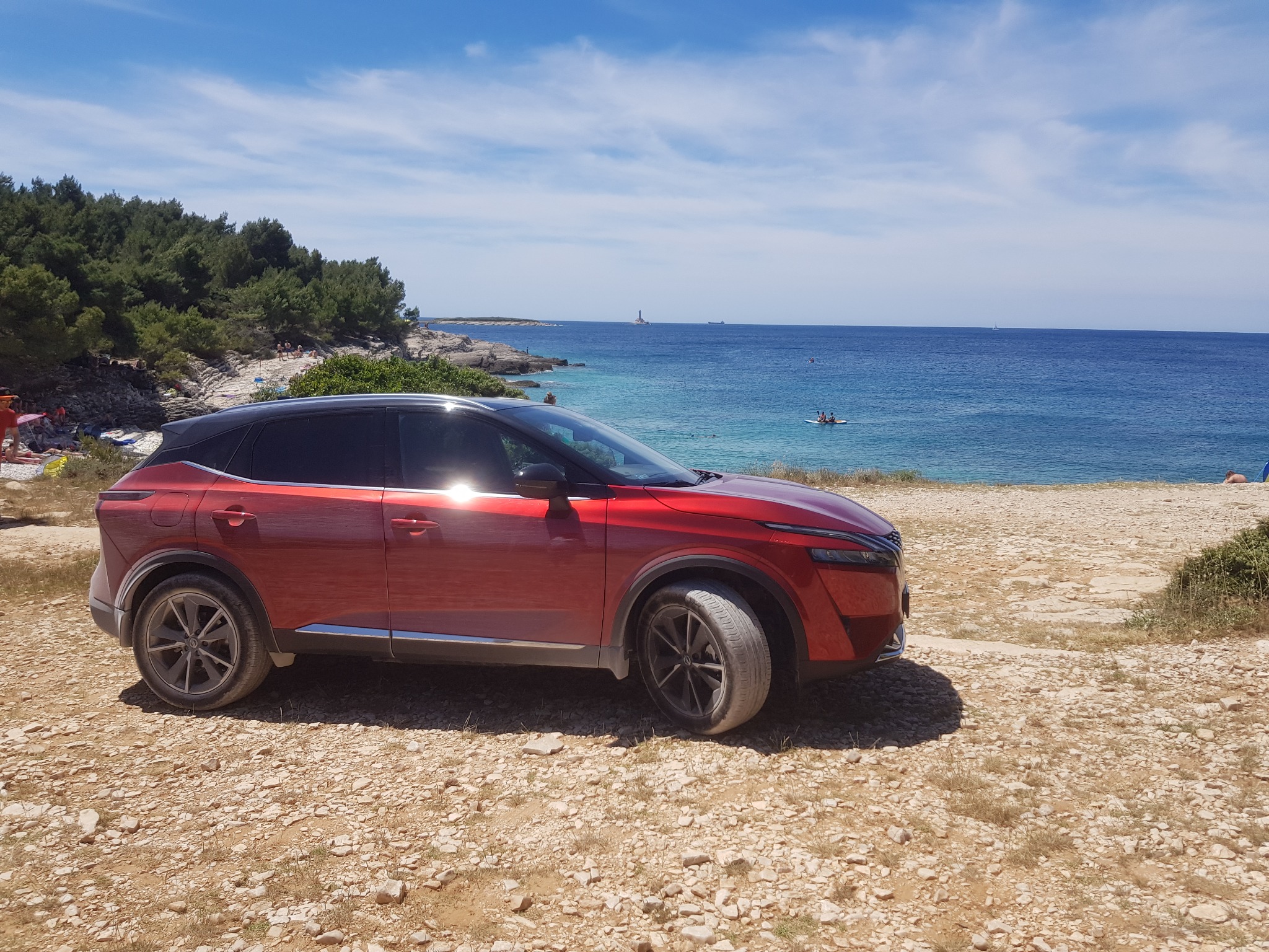 Czerwony SUV zaparkowany na kamienistej plaży z widokiem na błękitne morze i odległą latarnię morską.