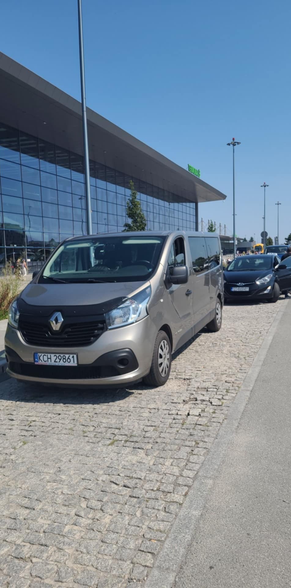 Szary bus Renault Trafic z czarnym grillem i osłoną na masce, zaparkowany na brukowanej nawierzchni przed nowoczesnym budynkiem z przeszkloną fasadą.