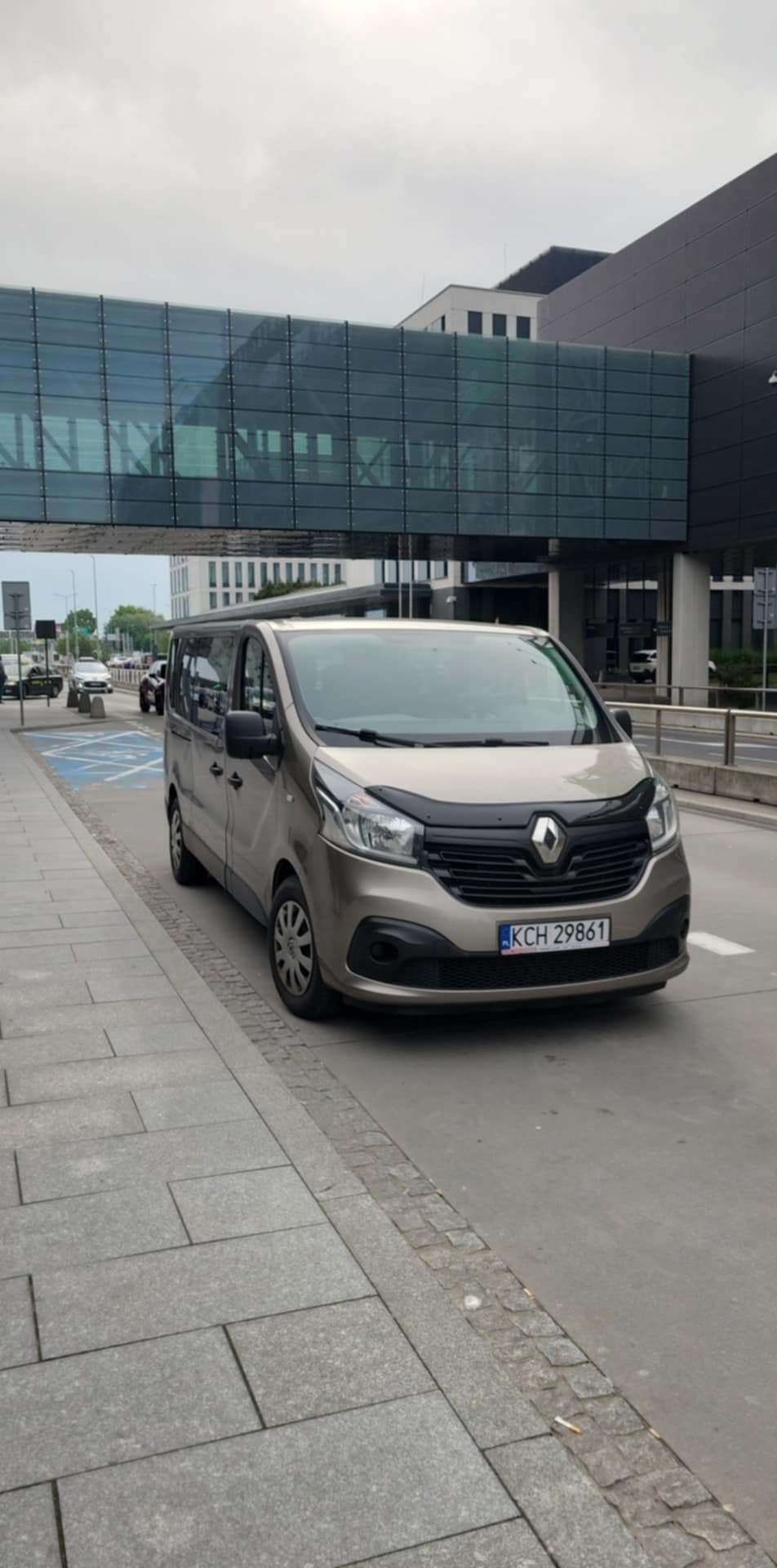 Beżowy van Renault z czarnym grillem i numerem KCH 29861 na tle nowoczesnego budynku z przeszklonym łącznikiem. Transport osobowy.