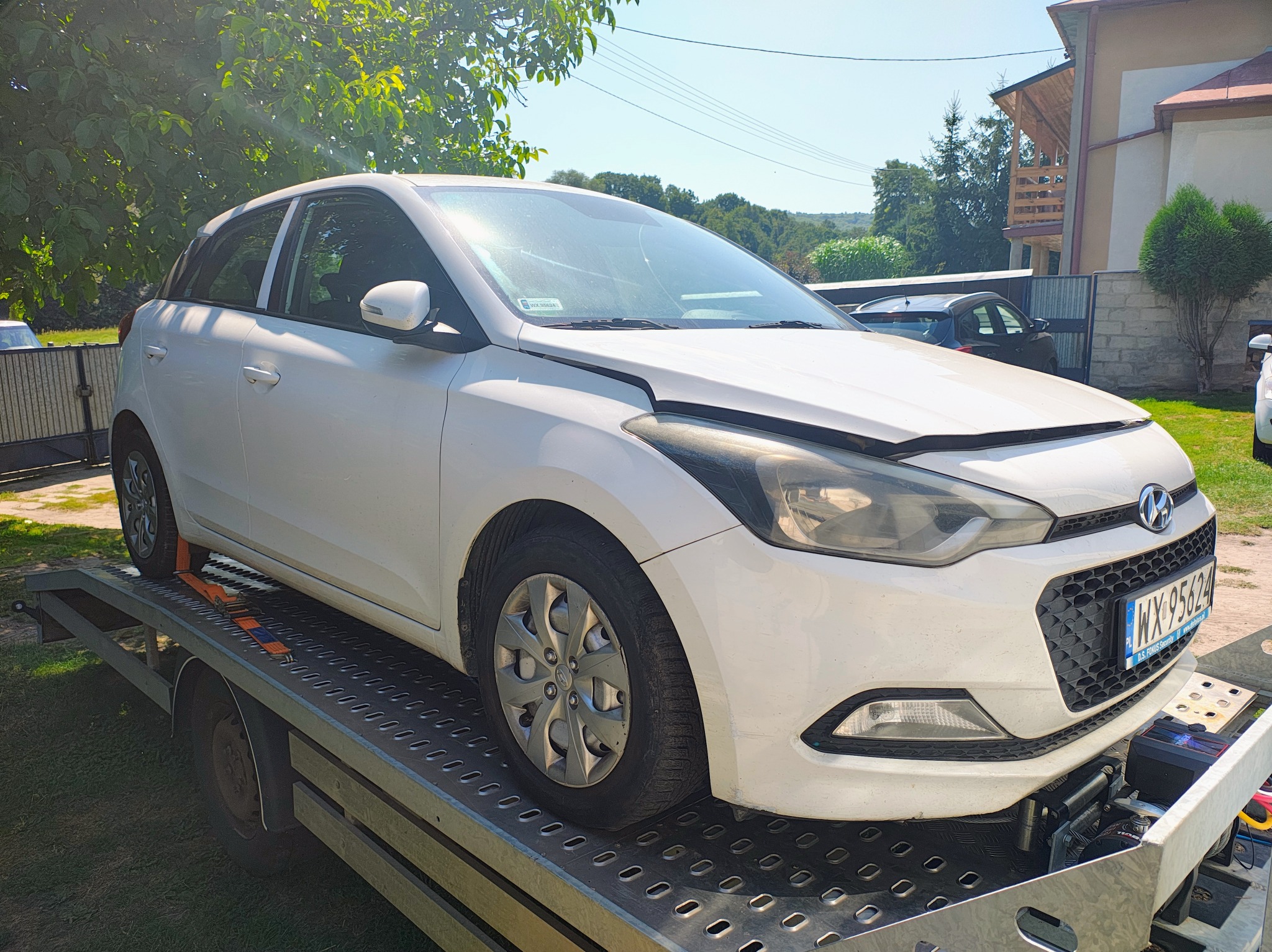 Biały samochód osobowy marki Hyundai z podniesioną maską, umieszczony na platformie lawety. Widoczny numer rejestracyjny. Tło stanowi budynek i zieleń.