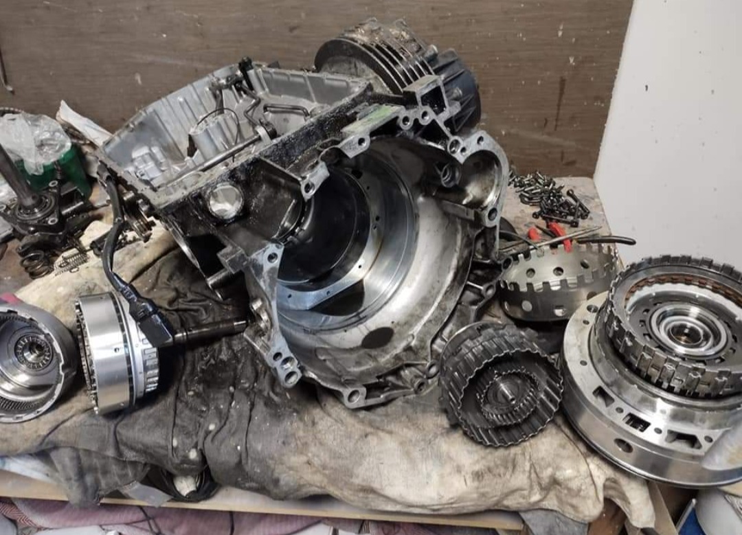 Rozłożona skrzynia biegów na warsztatowym stole, widoczne elementy przekładni i obudowa, ślady oleju i zabrudzeń. Narzędzia w tle sugerują etap naprawy.