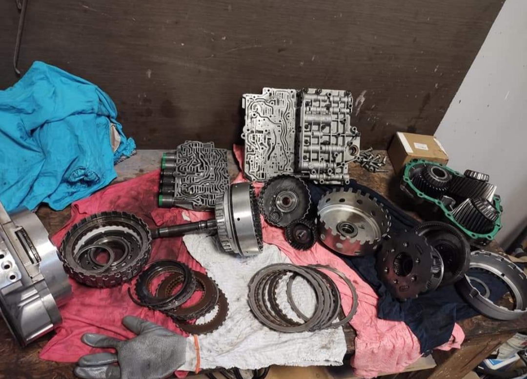 Rozłożona skrzynia biegów na stole warsztatowym, widoczne elementy przekładni, sterowniki i inne podzespoły. Różne rozmiary i kształty części, ślady oleju i smaru.