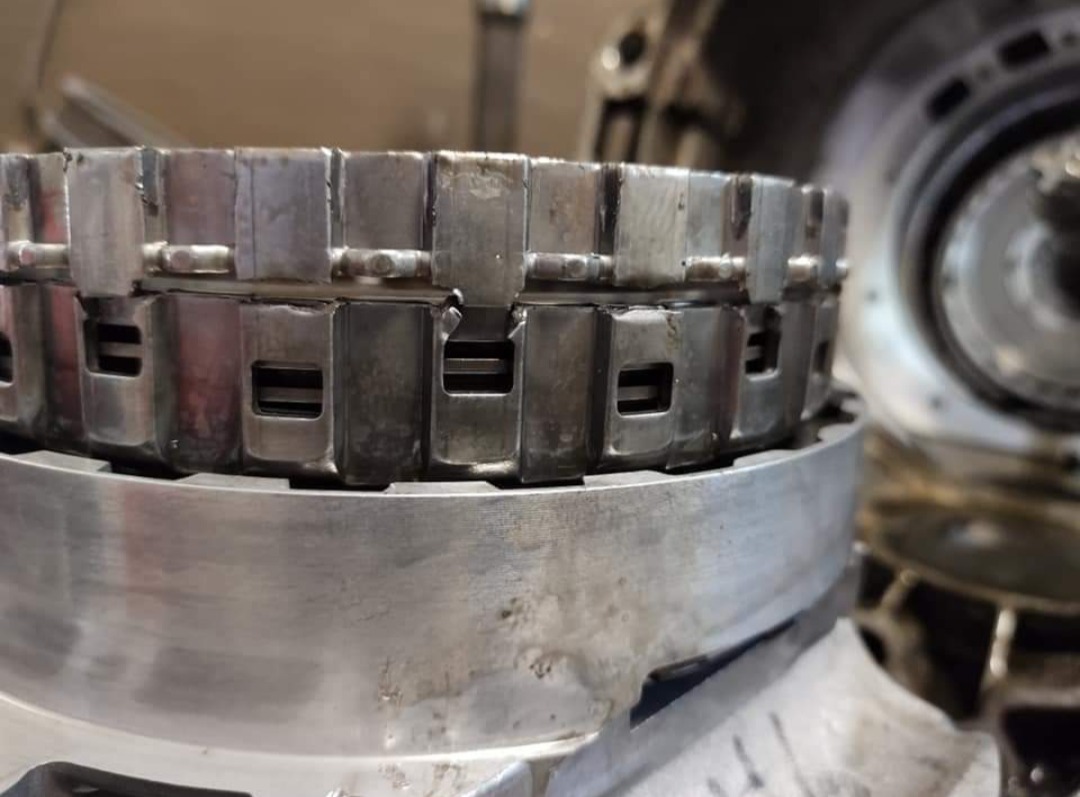 Metalowa część samochodowa z widocznymi śladami użytkowania, prawdopodobnie element skrzyni biegów, sfotografowana w warsztacie samochodowym.