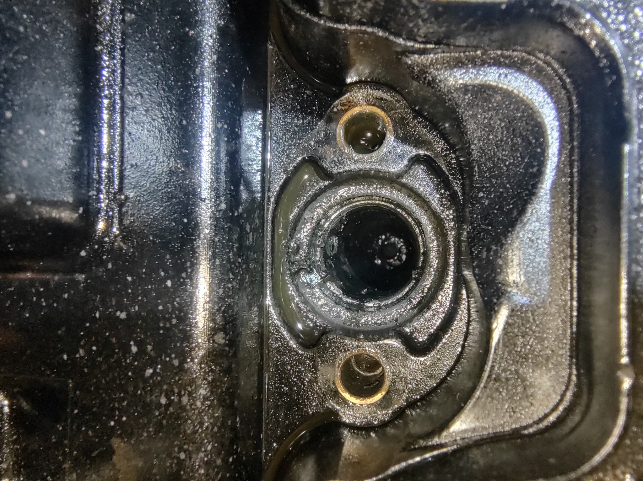 Zbliżenie na fragment silnika samochodowego z widocznymi otworami montażowymi i śladami oleju, ukazujące detale konstrukcyjne i zużycie eksploatacyjne.