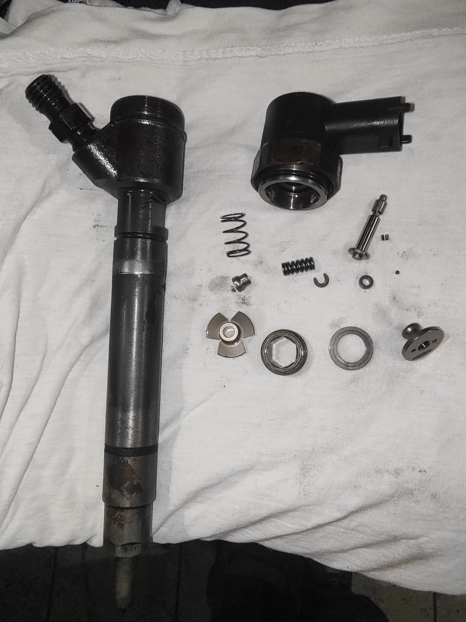 Rozłożony na części wtryskiwacz paliwa na białej szmatce. Widoczne elementy składowe, sprężyny, uszczelki i korpus wtryskiwacza. Precyzyjny demontaż.